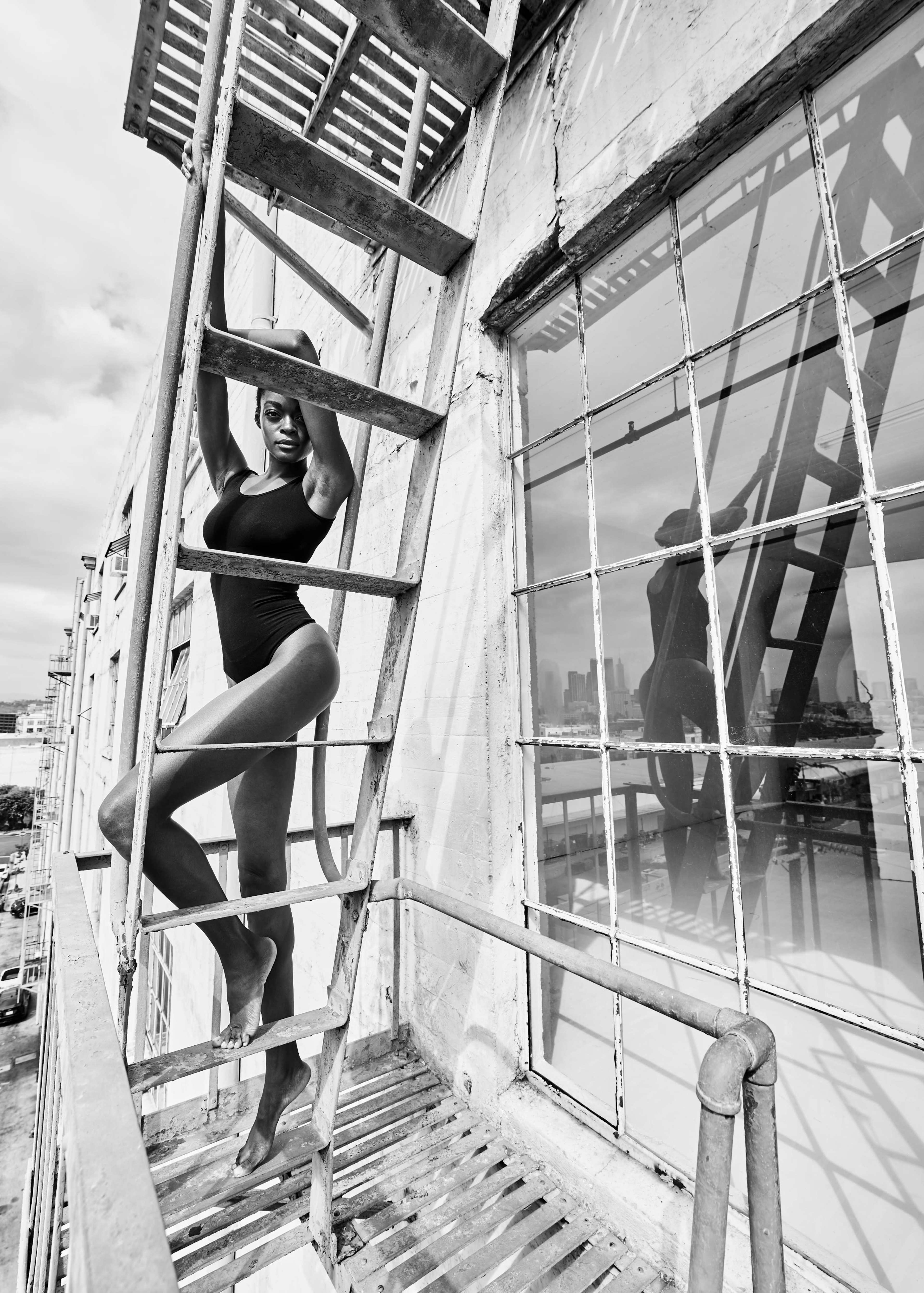 A dancer poses on a fire escape outside a building, reflecting in the large window beside her.