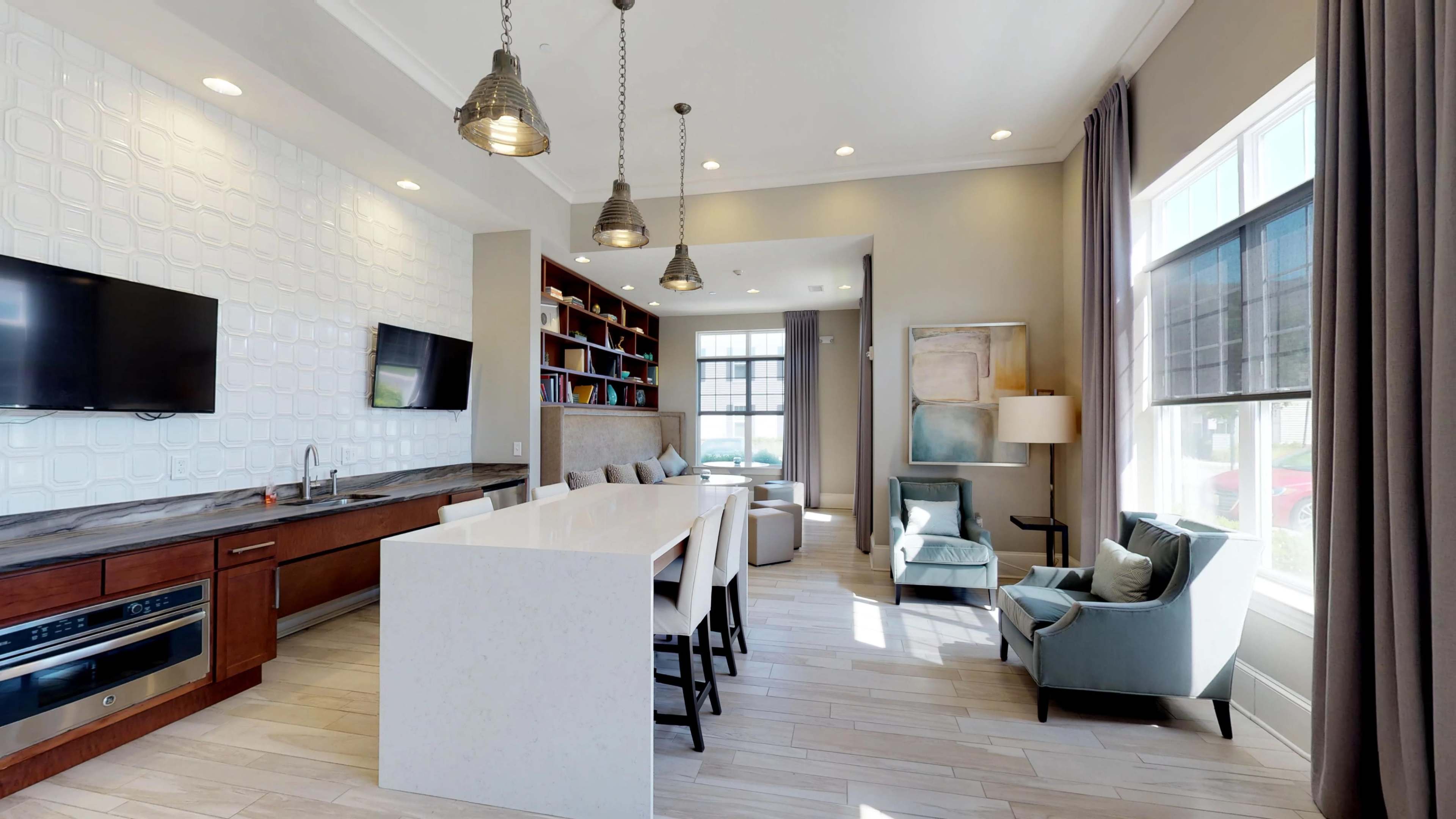 A modern interior space featuring a kitchen area with a large white countertop, seating, bookshelf, and a cozy seating arrangement near windows.