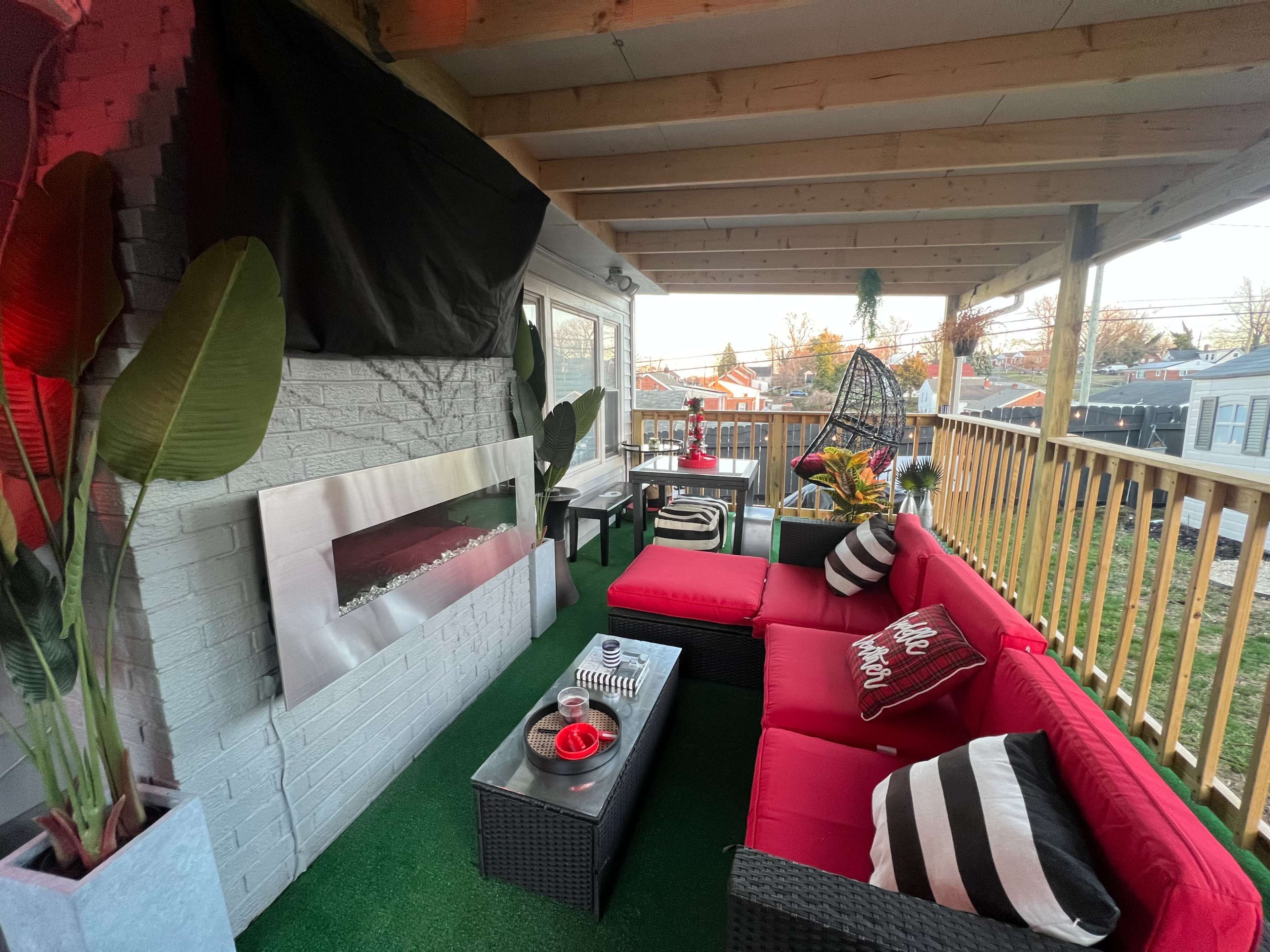 A covered outdoor seating area featuring red couches, a glass coffee table, and a wall-mounted fireplace.