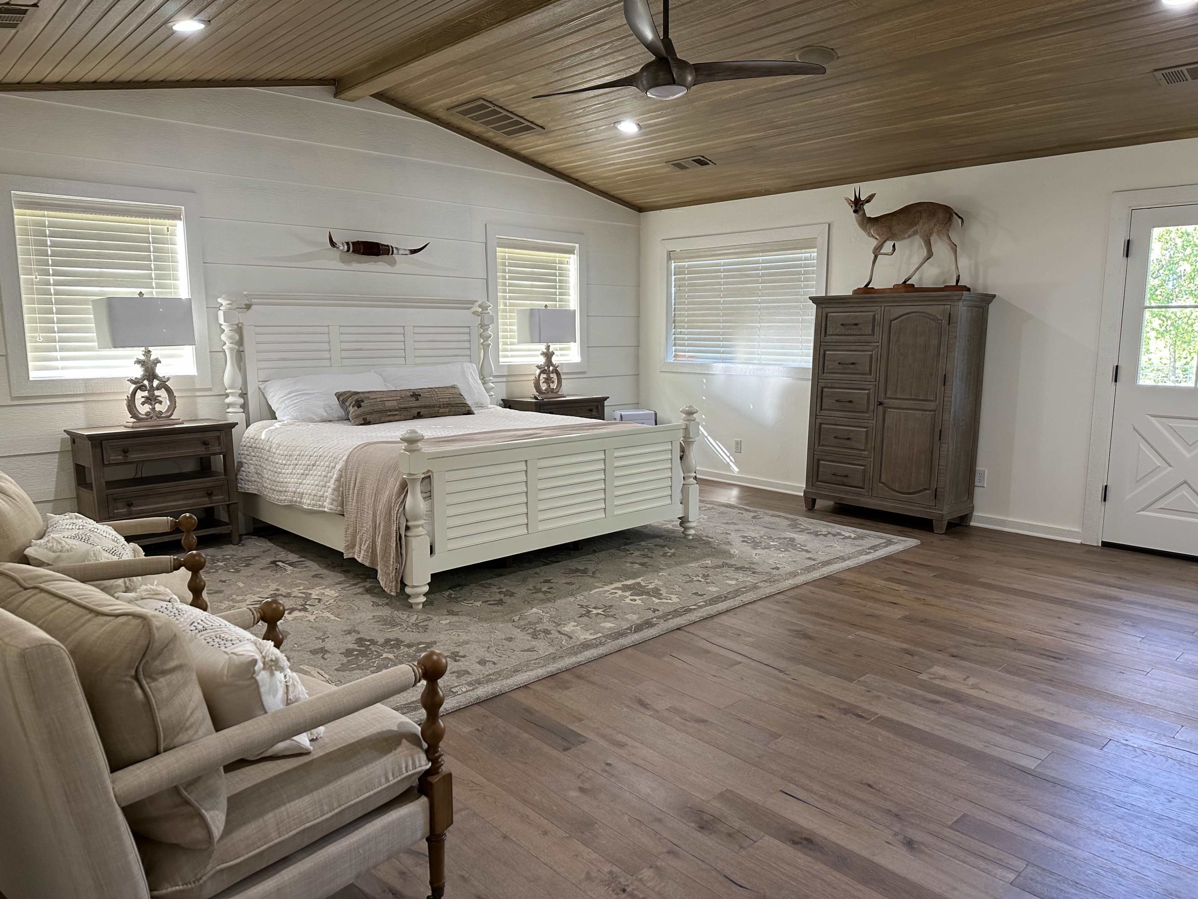 A spacious bedroom with a white wooden bed, beige bedding, a pair of lamps on nightstands, a wooden dresser, and a chair set in a cozy layout.