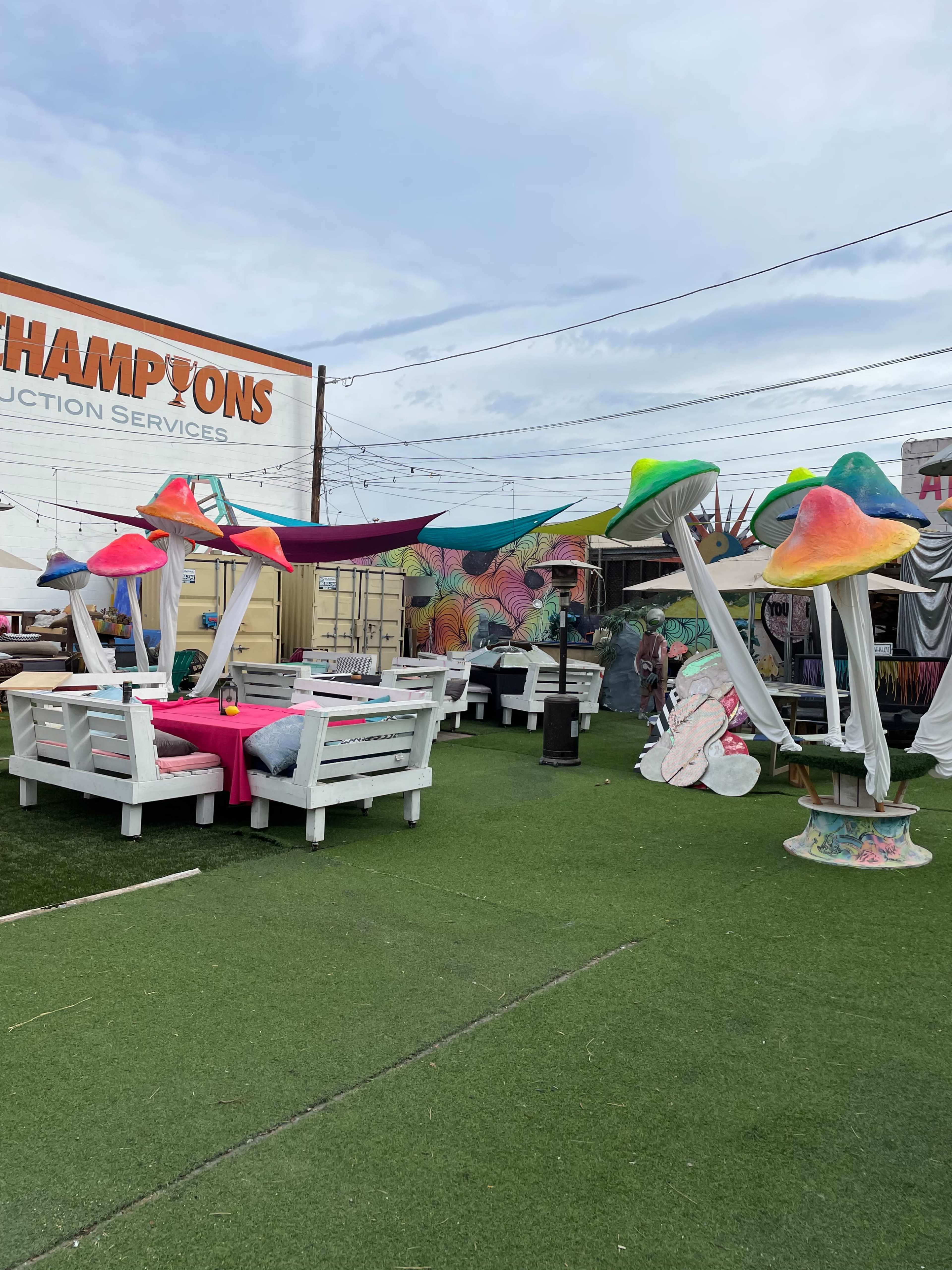 The scene features a colorful outdoor area with oversized mushroom sculptures, white furniture, and a vibrant mural in the background.