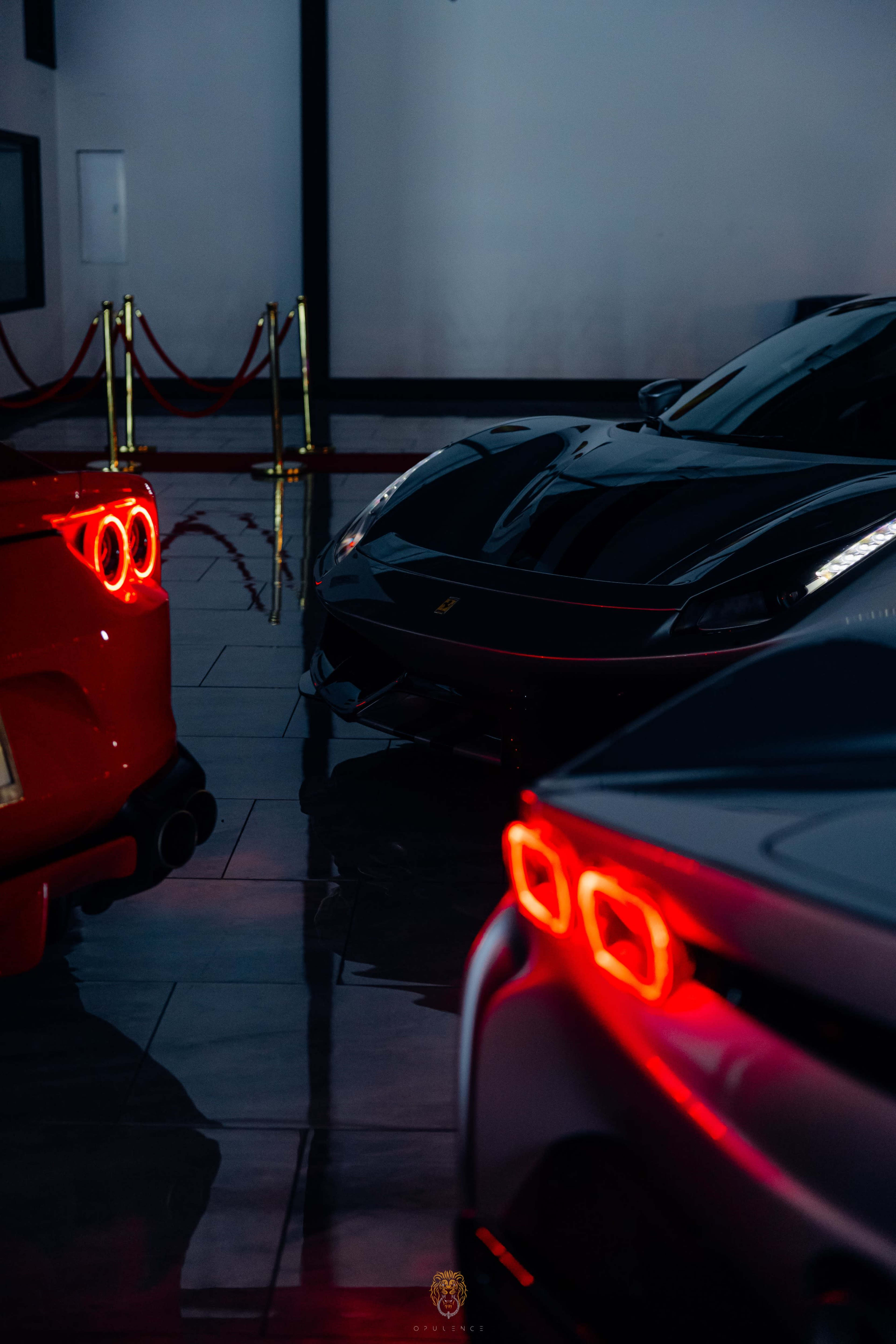 Three luxury sports cars are parked in a showroom, with their rear lights glowing red against a polished floor.