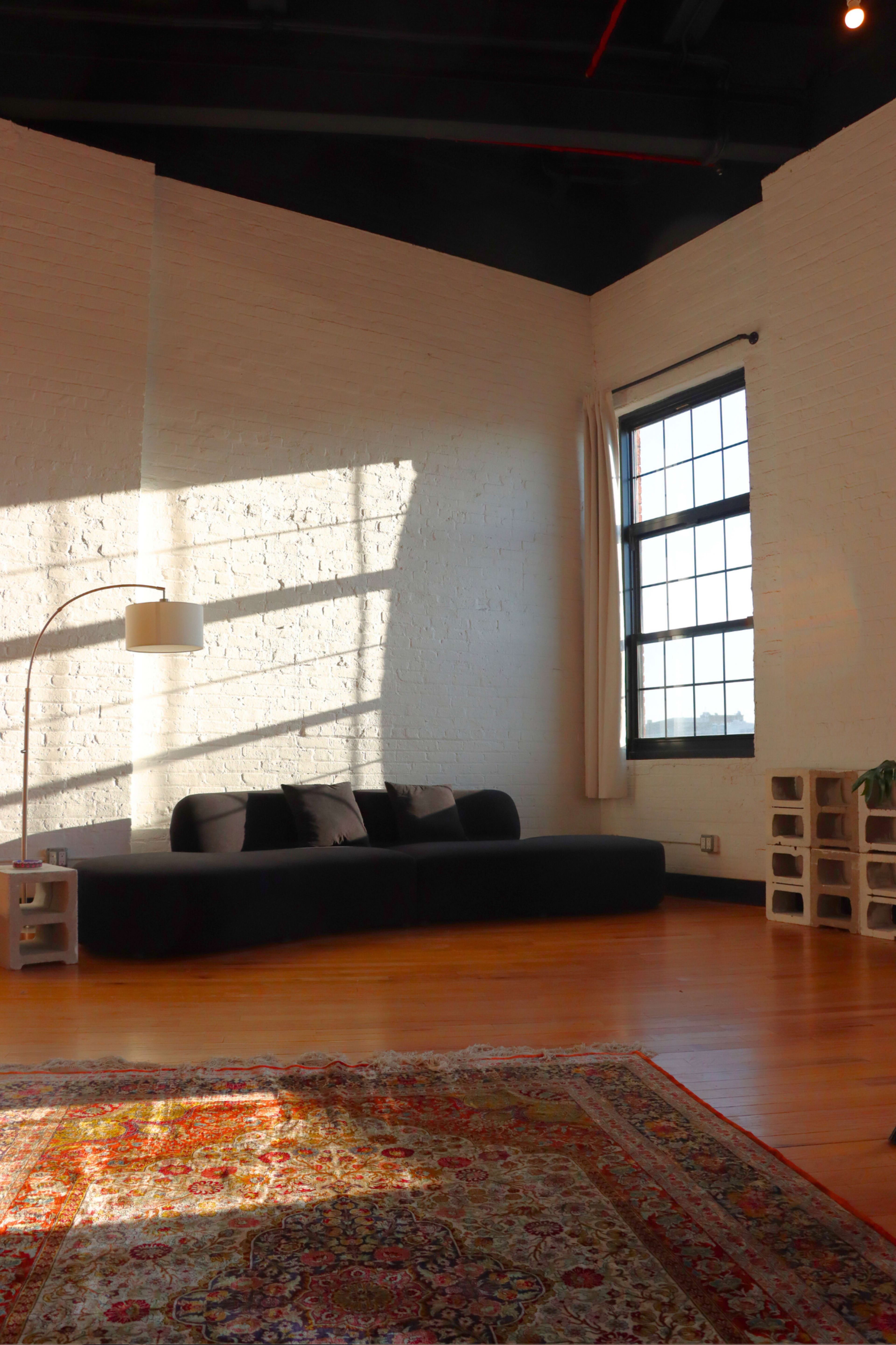 The image shows a modern living space with a sectional sofa, a floor lamp, a large window, and a patterned rug on wooden flooring.