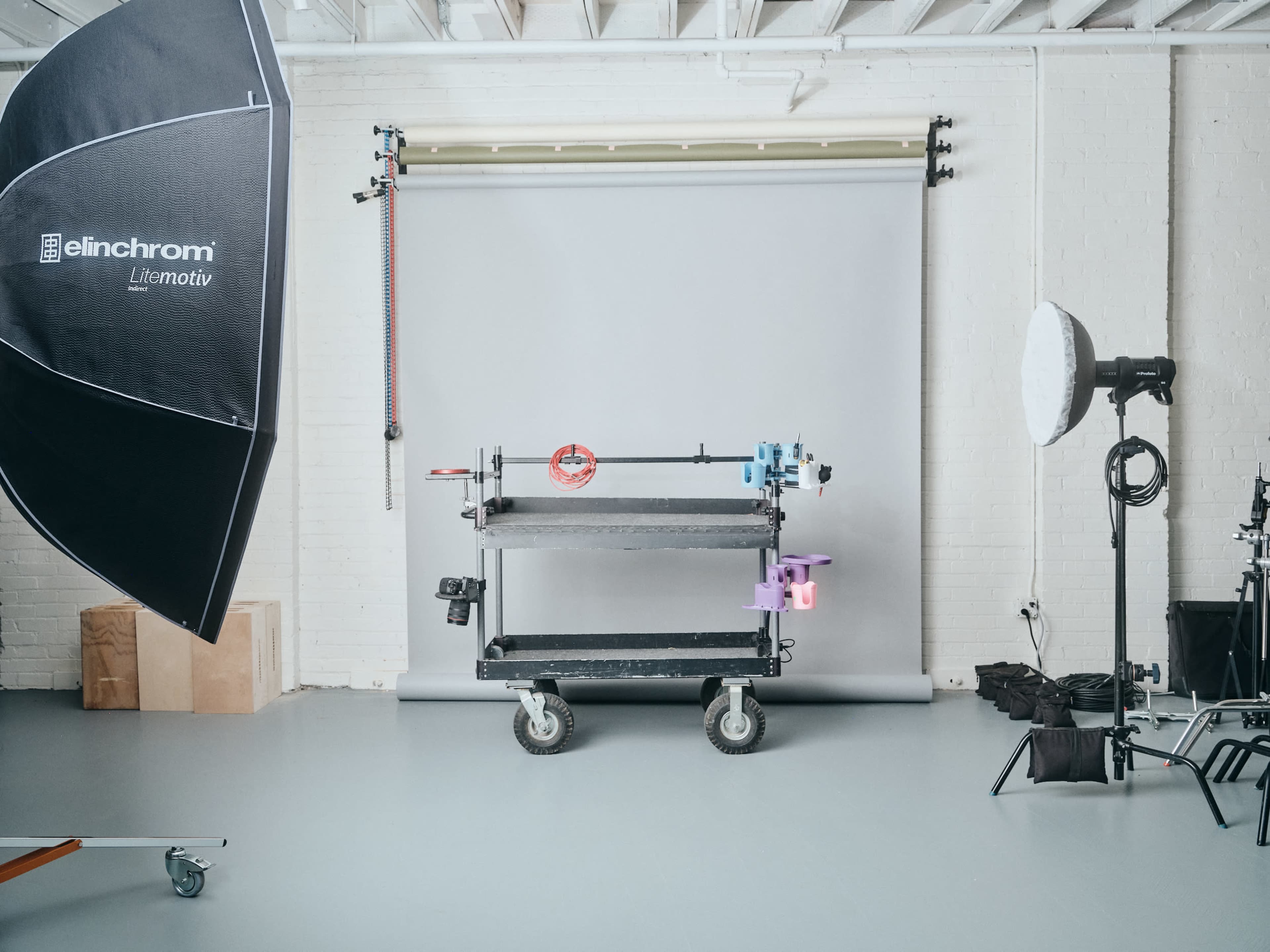 A photography studio features a light setup with a softbox and an umbrella, alongside a wheeled cart holding various equipment against a gray backdrop.