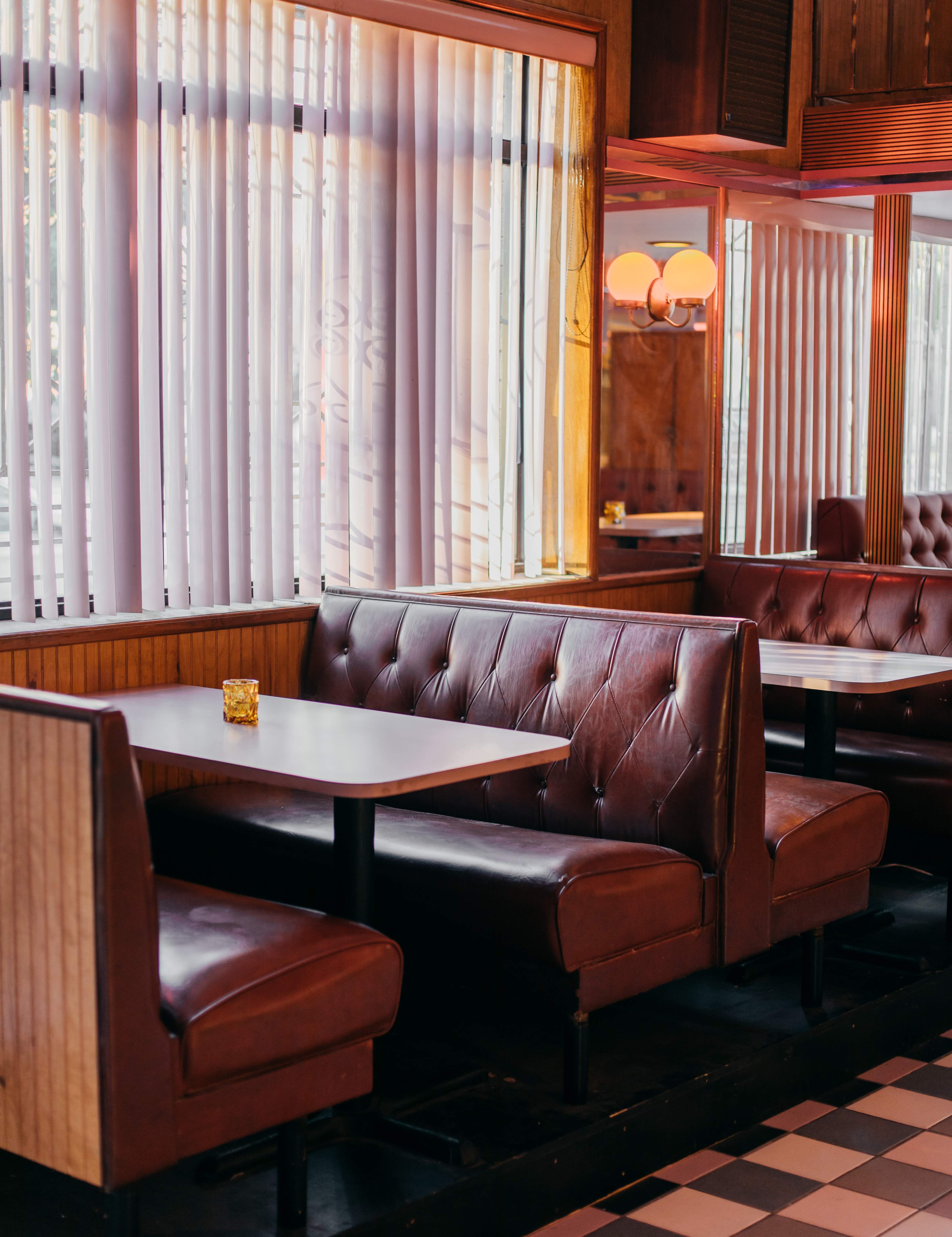 A cozy diner interior featuring brown leather booths arranged around white-topped tables, with large windows adorned by vertical blinds.
