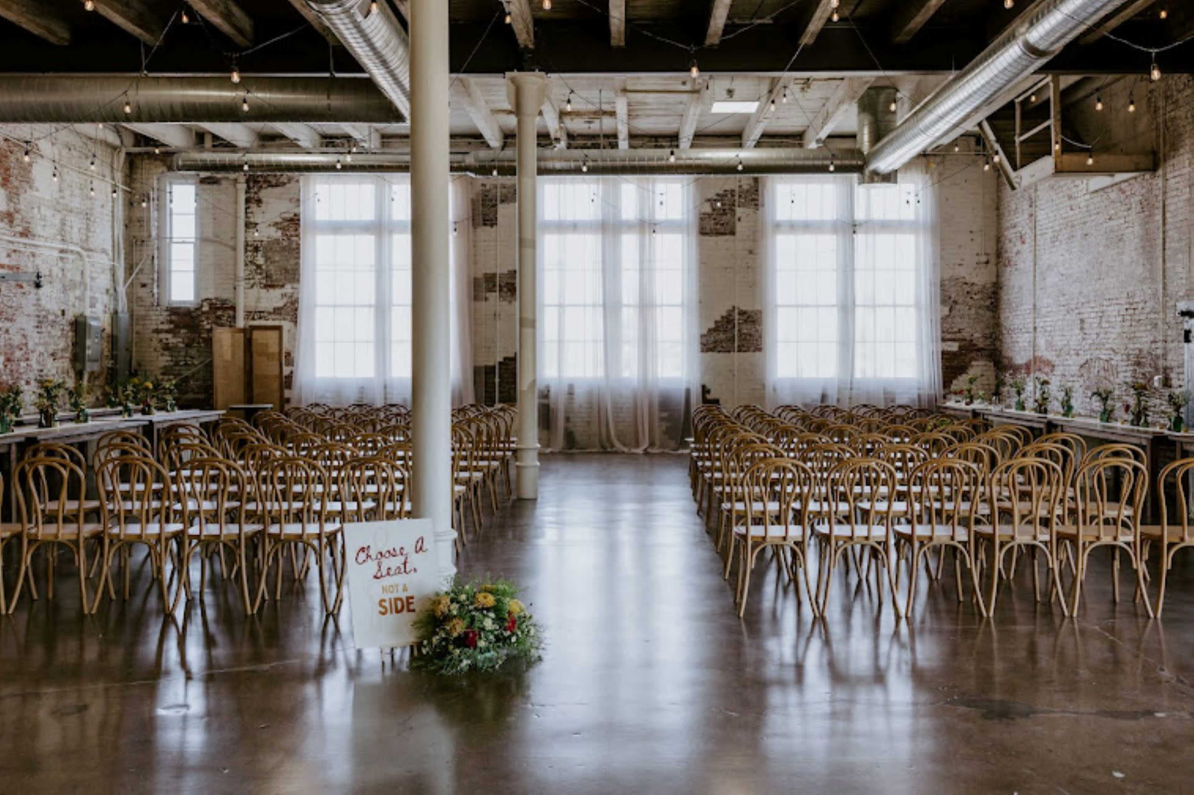 A spacious event hall features rows of tables and chairs arranged for a gathering, with large windows allowing natural light to fill the room.