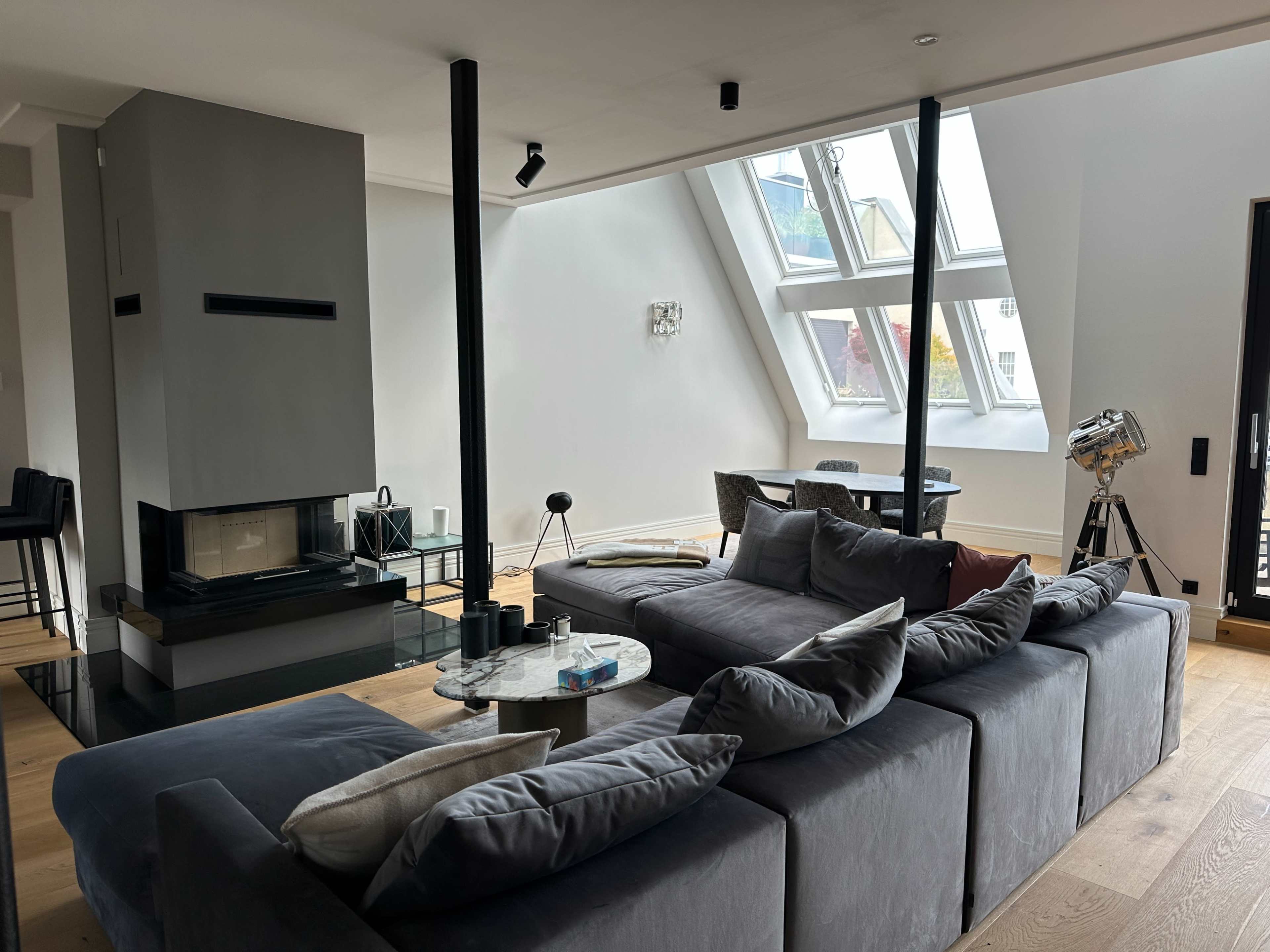 The image shows a modern living room with a gray sectional sofa, a marble coffee table, and a contemporary fireplace, featuring large windows that allow natural light to enter the space.
