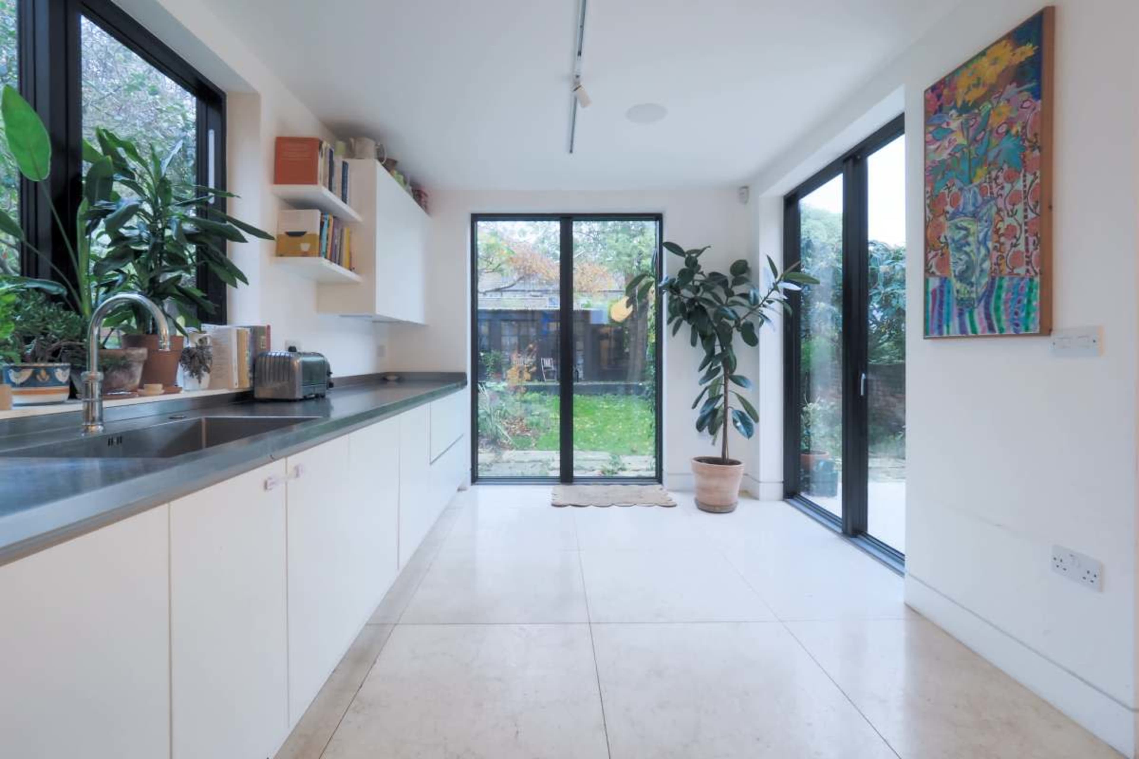 A bright kitchen with a long counter, large windows, and potted plants opens to a garden view.