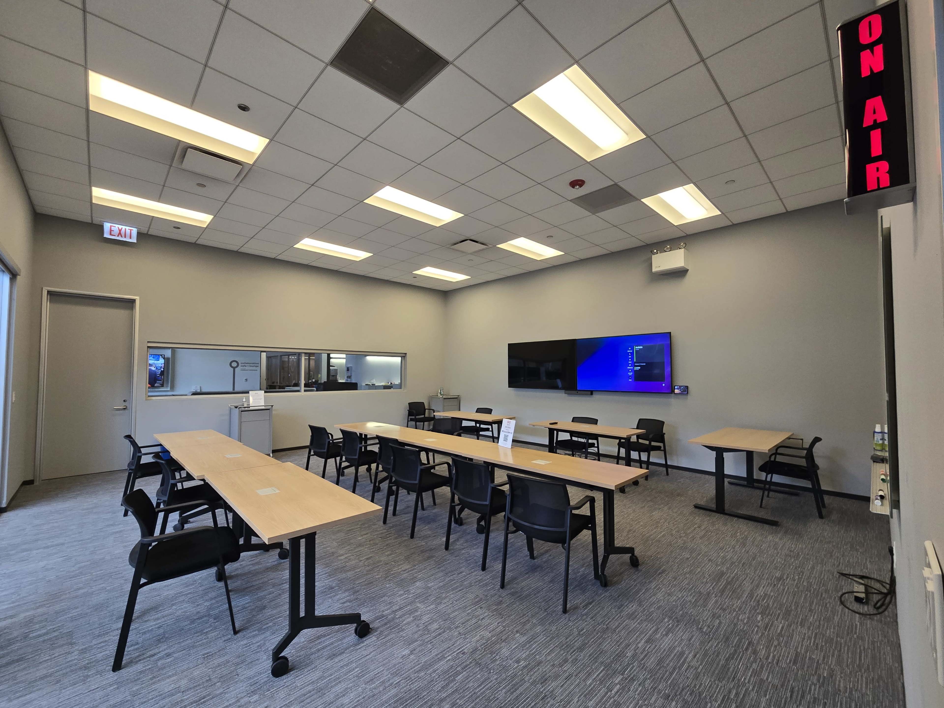 The image shows a modern conference room with multiple tables, chairs, a large screen on one wall, and an "ON AIR" sign.