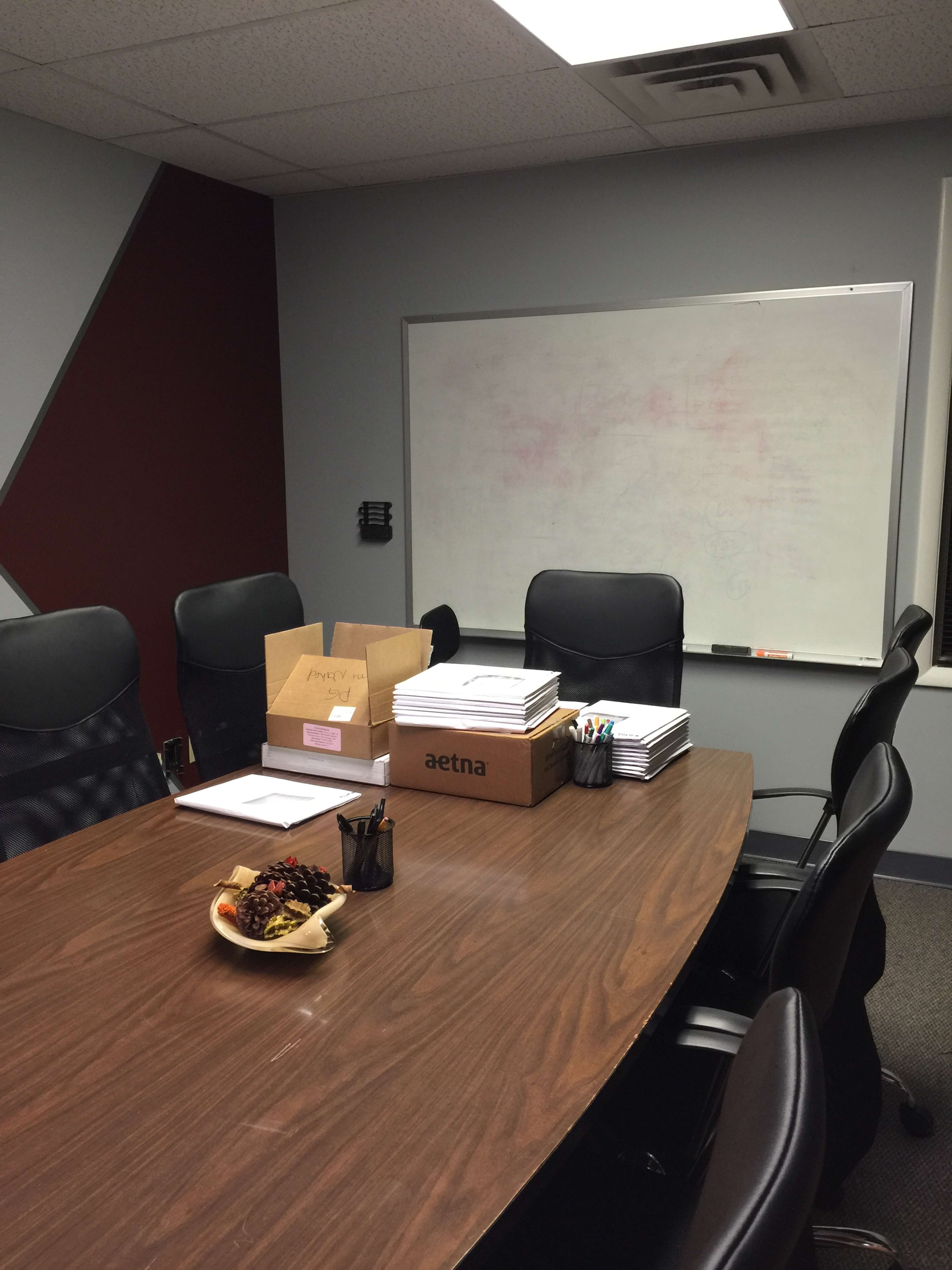 A conference room with a large wooden table surrounded by black chairs, featuring a whiteboard, a box labeled "aetna," and documents neatly stacked.