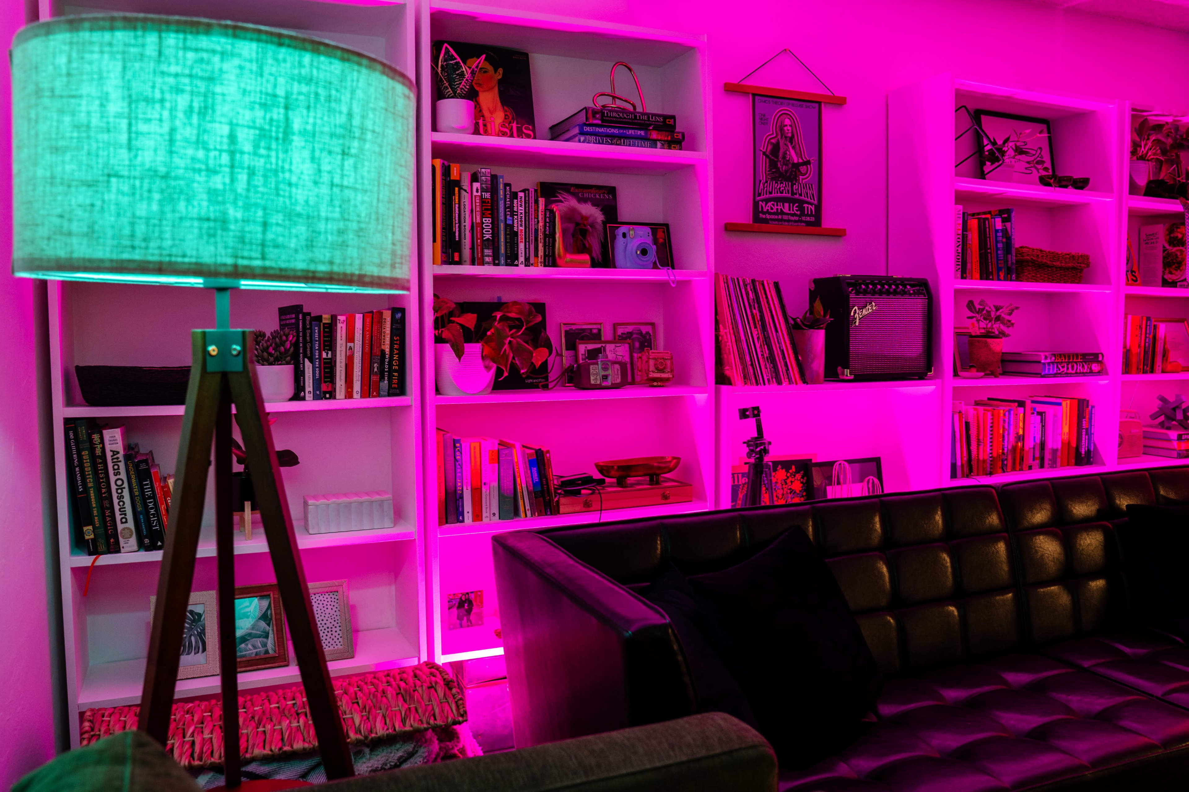 A modern living room features a green tripod lamp beside a black leather sofa, with pink ambient lighting highlighting a shelf filled with books and decorative items.
