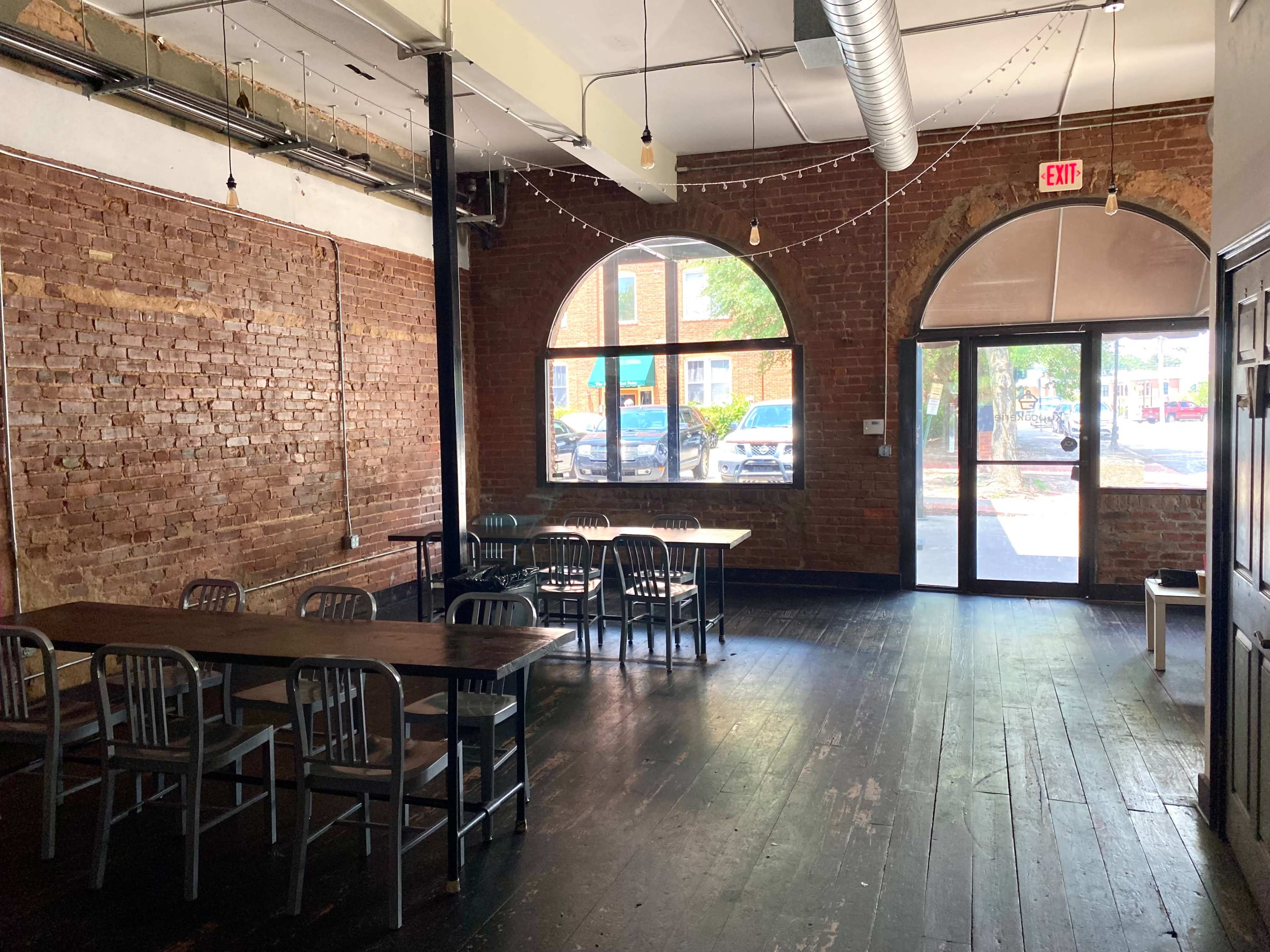 A spacious interior with exposed brick walls, wooden floors, and several tables and chairs arranged near large arched windows.