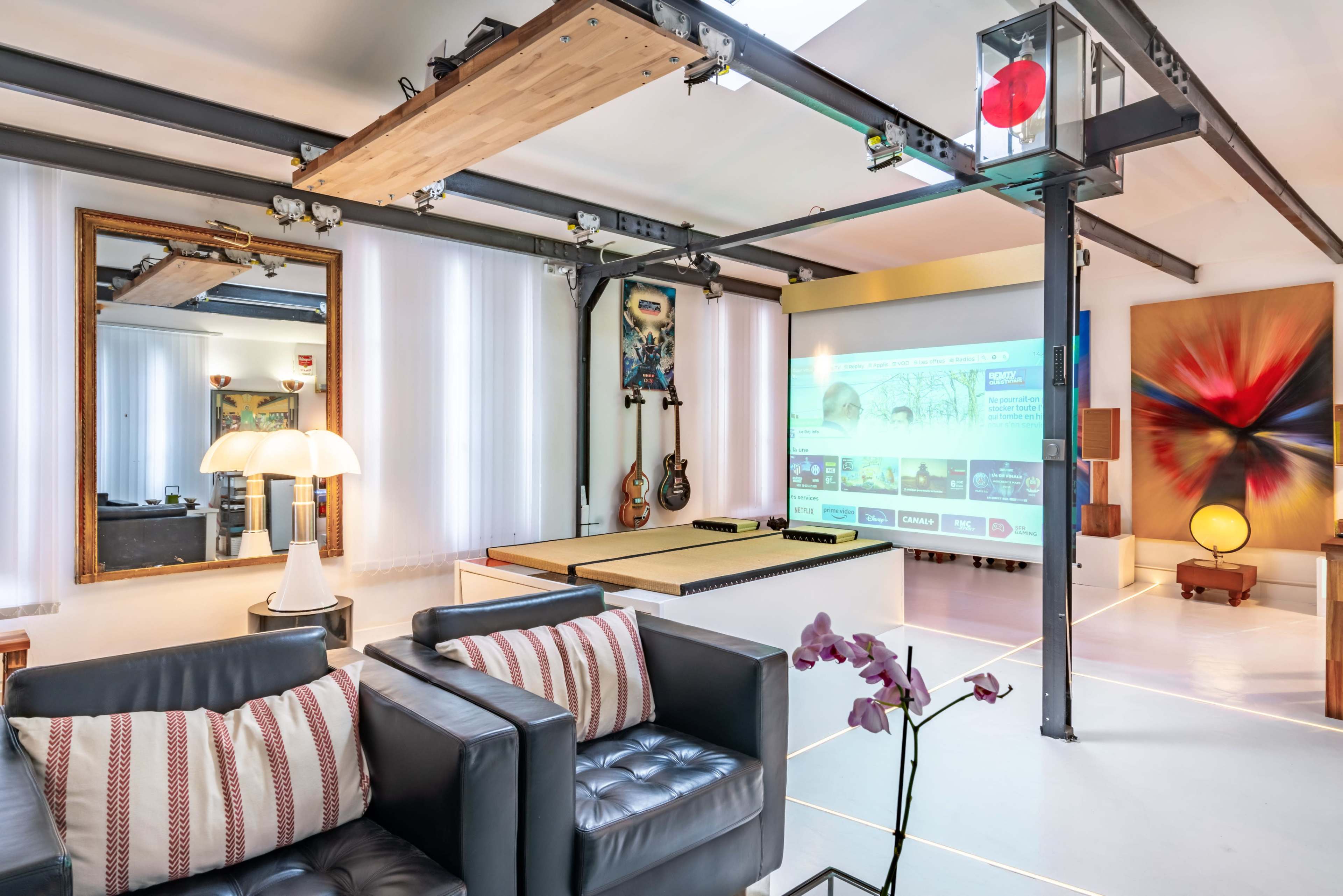 The image shows a modern, stylish living space featuring a projector screen, a seating area with leather sofas, artwork on the walls, and large windows providing natural light.