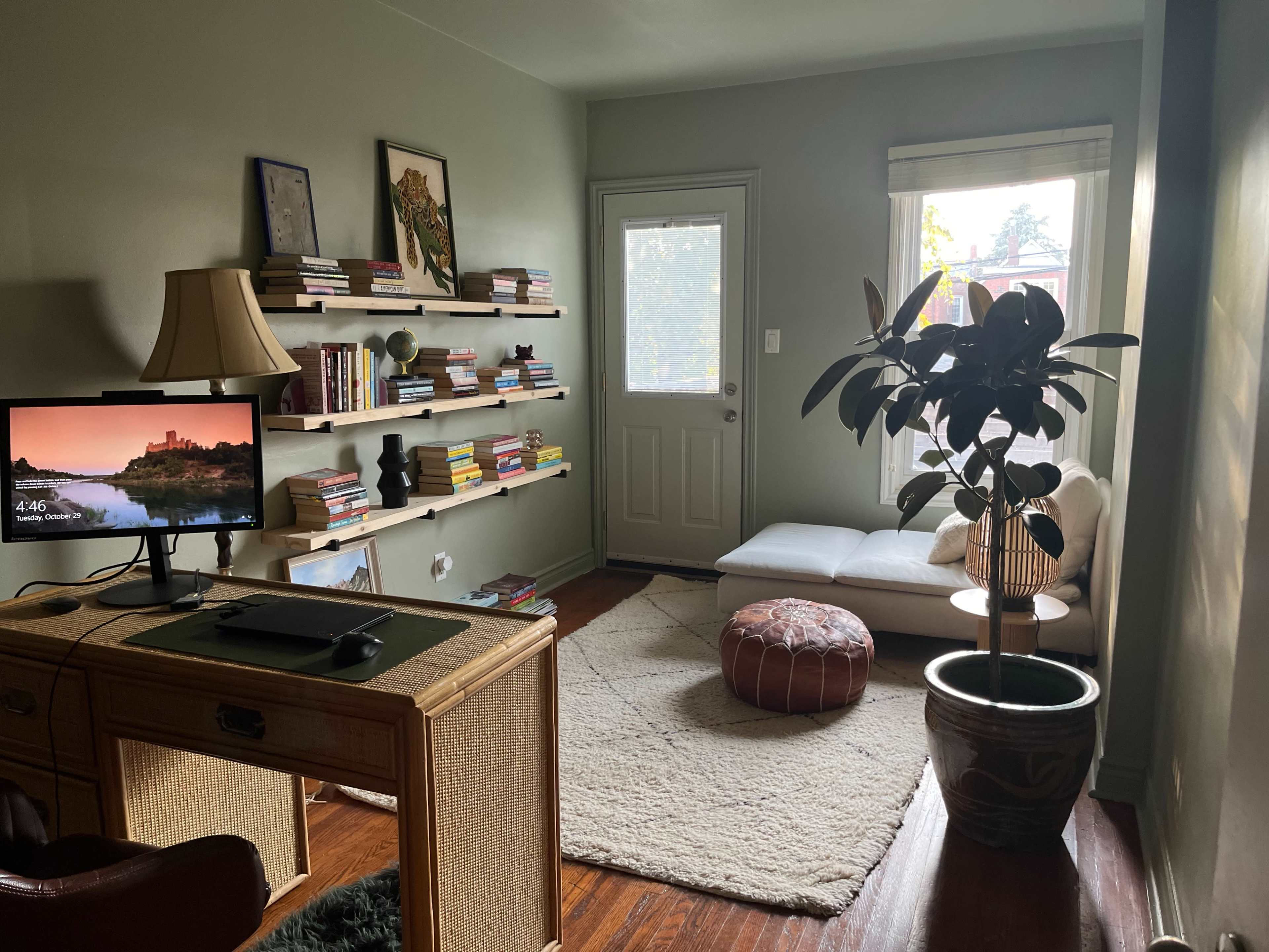 The image shows a cozy room with a small desk, bookshelves filled with books, a door leading outside, a chaise lounge, a round ottoman, and a potted plant.