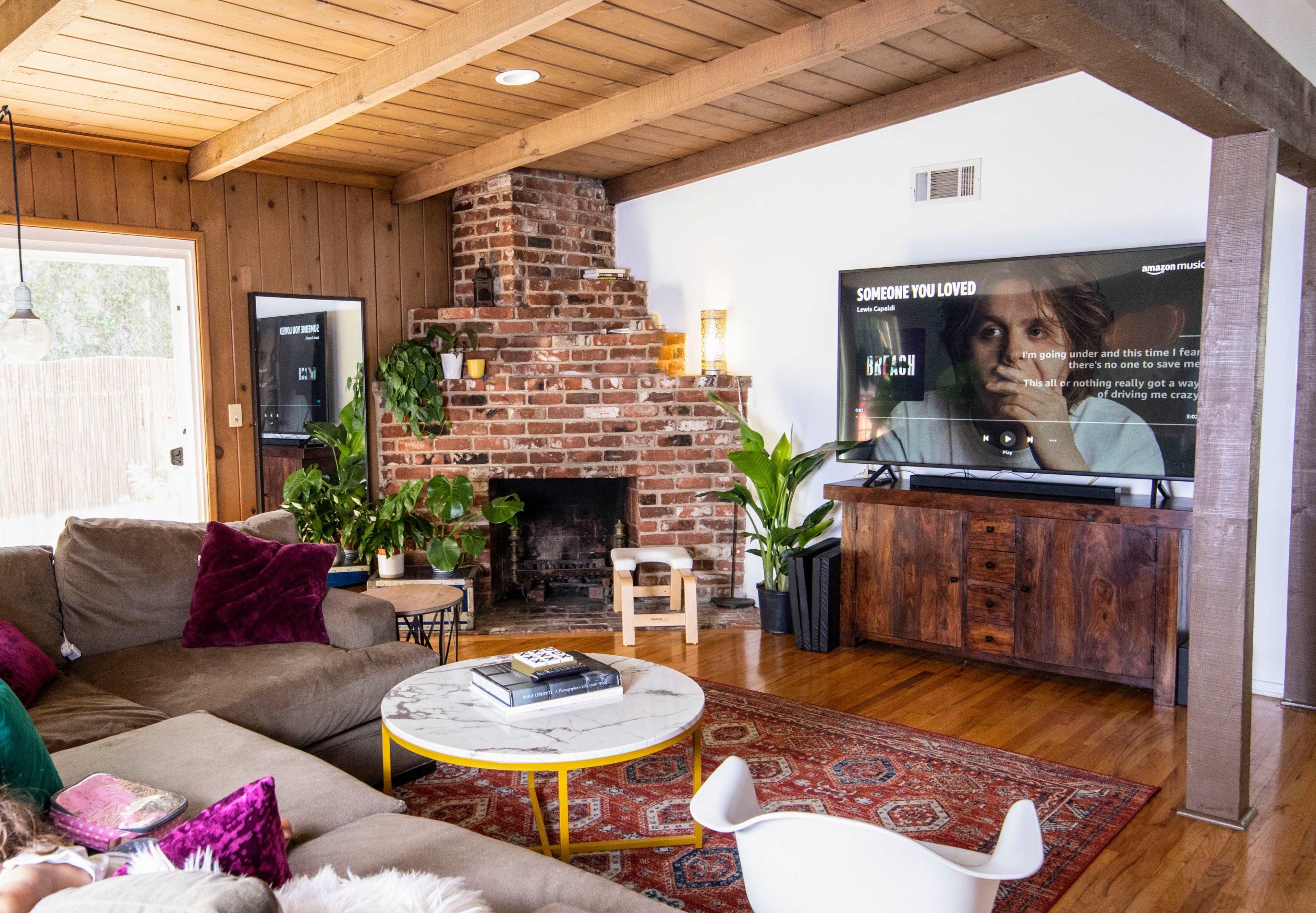 A cozy living room features a stone fireplace, a large television displaying a streaming service, and a sofa arranged around a coffee table with plants and decorative items.