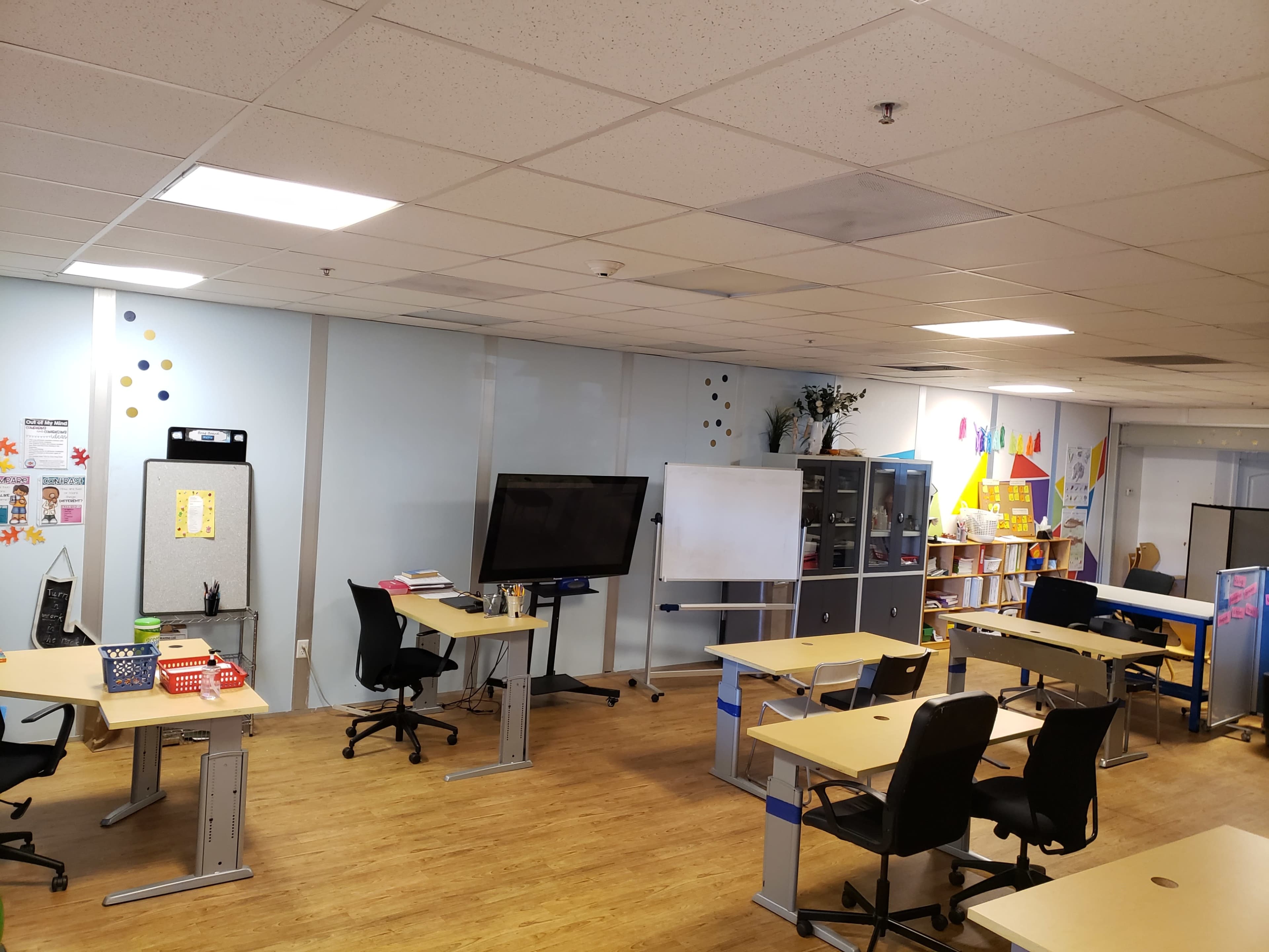 A modern classroom with several desks, a whiteboard, a TV screen, and shelves filled with educational materials.