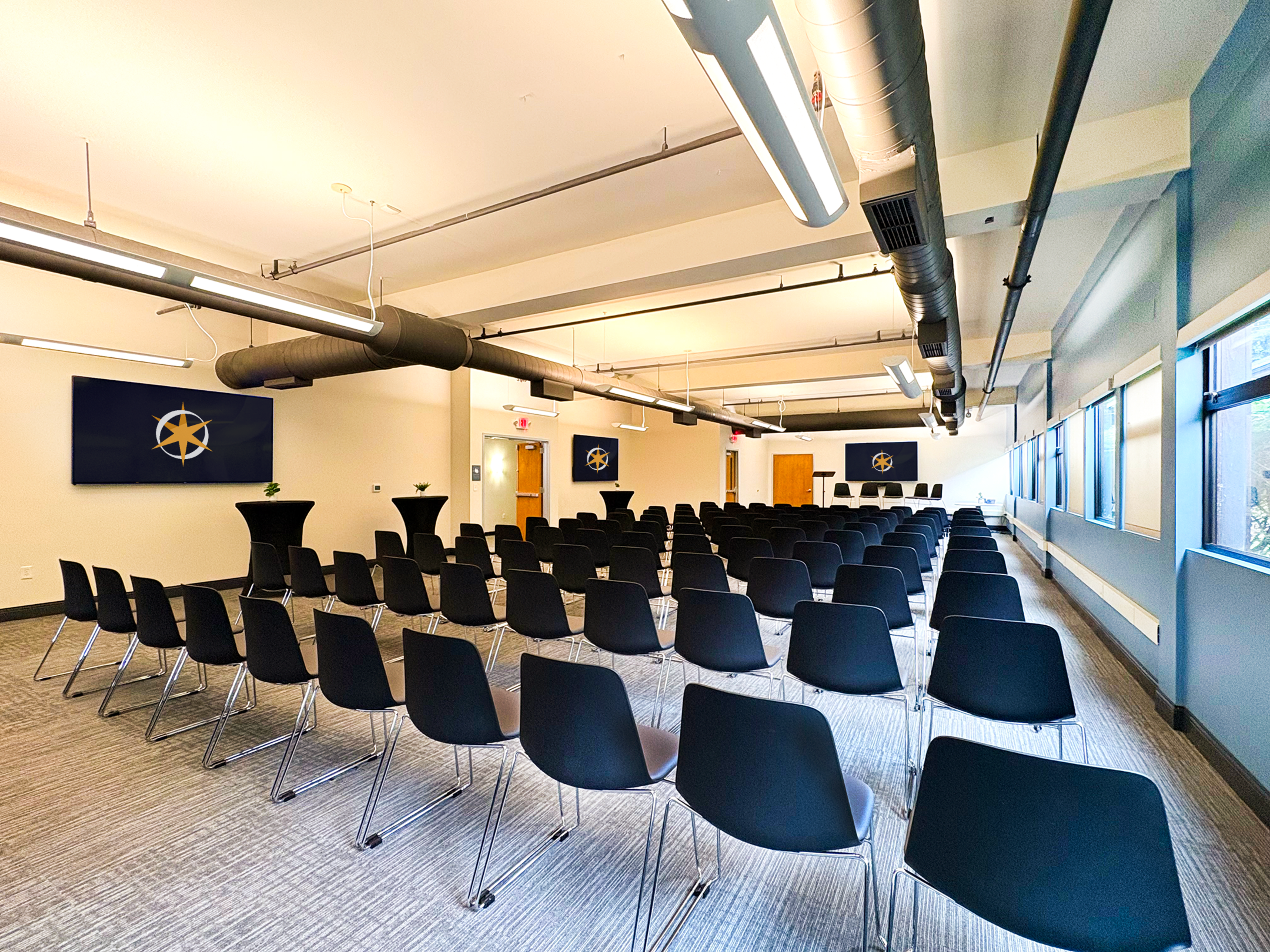 The image shows a spacious conference room arranged with rows of black chairs facing a podium and two screens on the front wall.