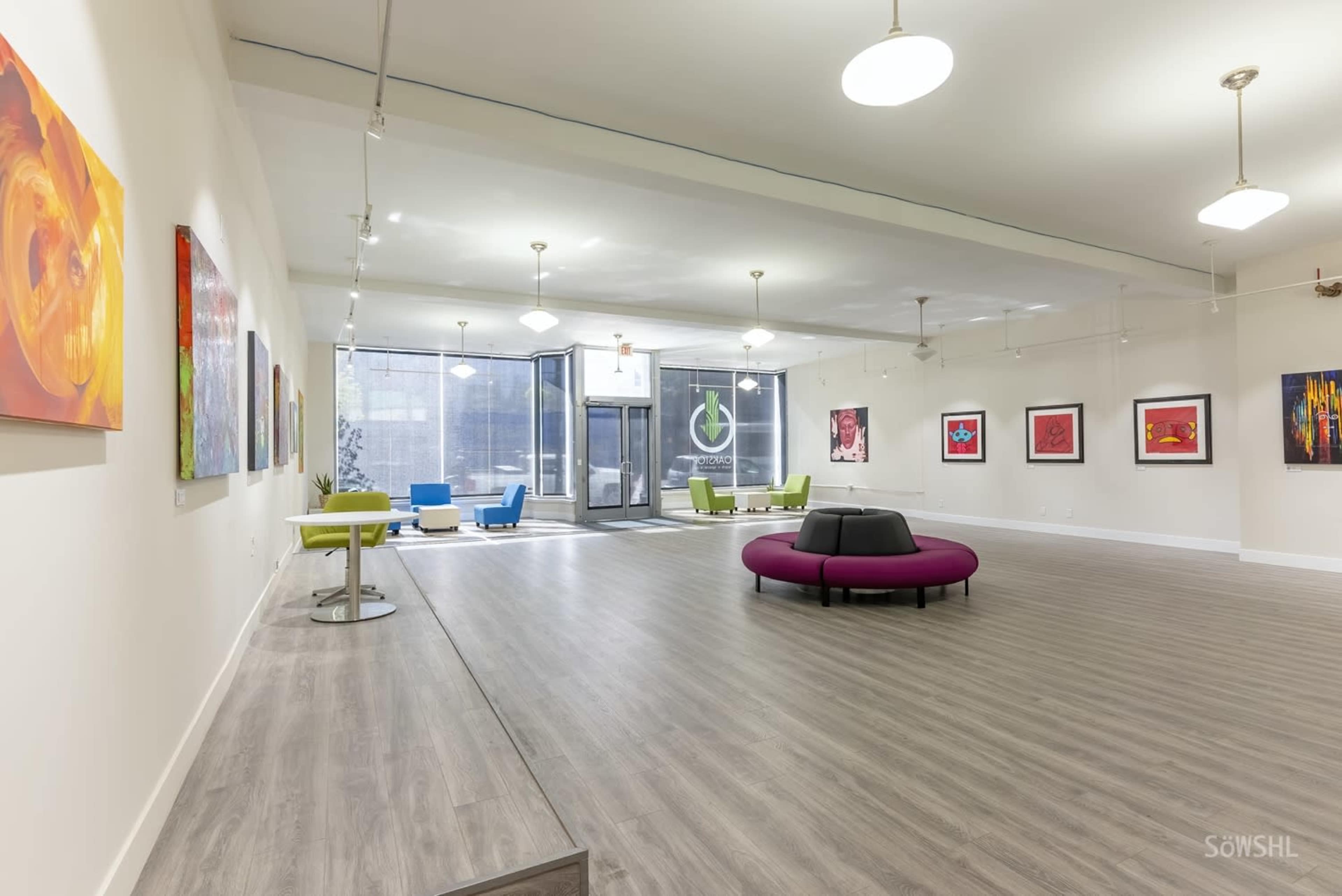 A spacious art gallery with colorful paintings on white walls, modern seating in the center, and large windows letting in natural light.