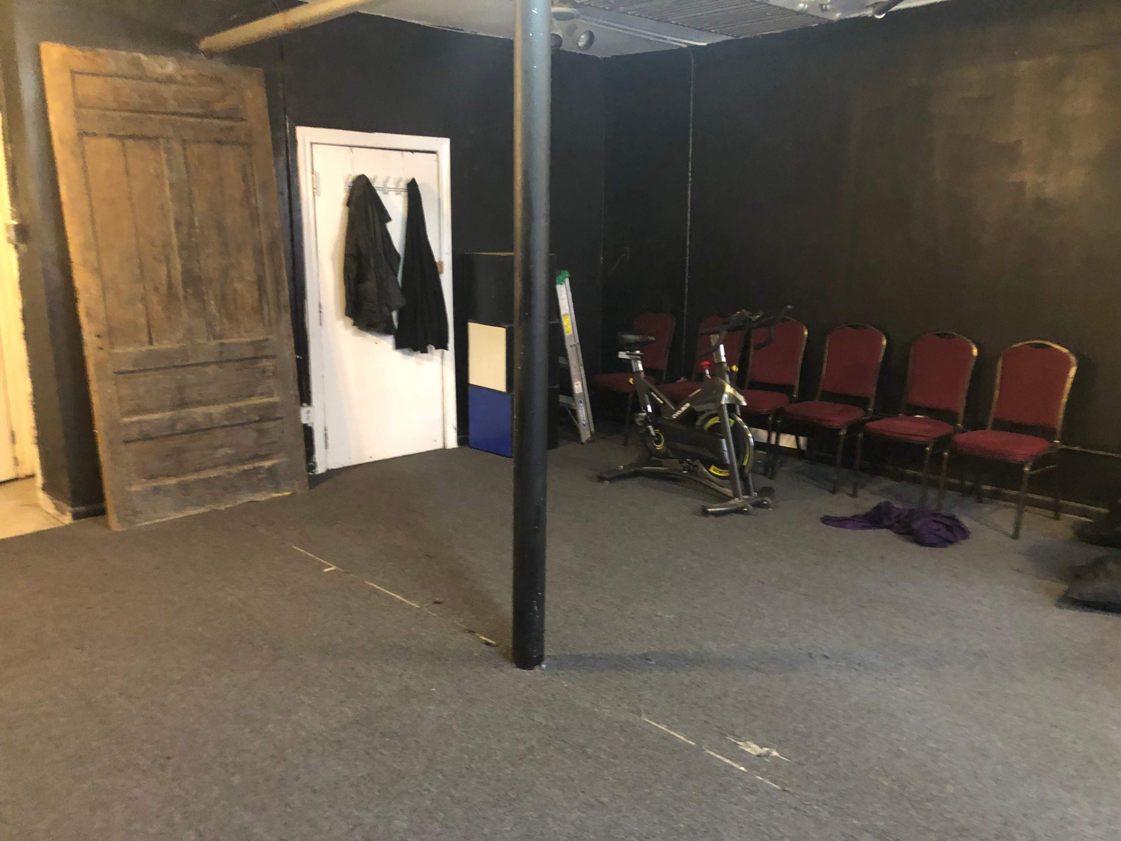 A dimly lit room with dark walls, a wooden door, a bike, and several red chairs arranged against one wall.