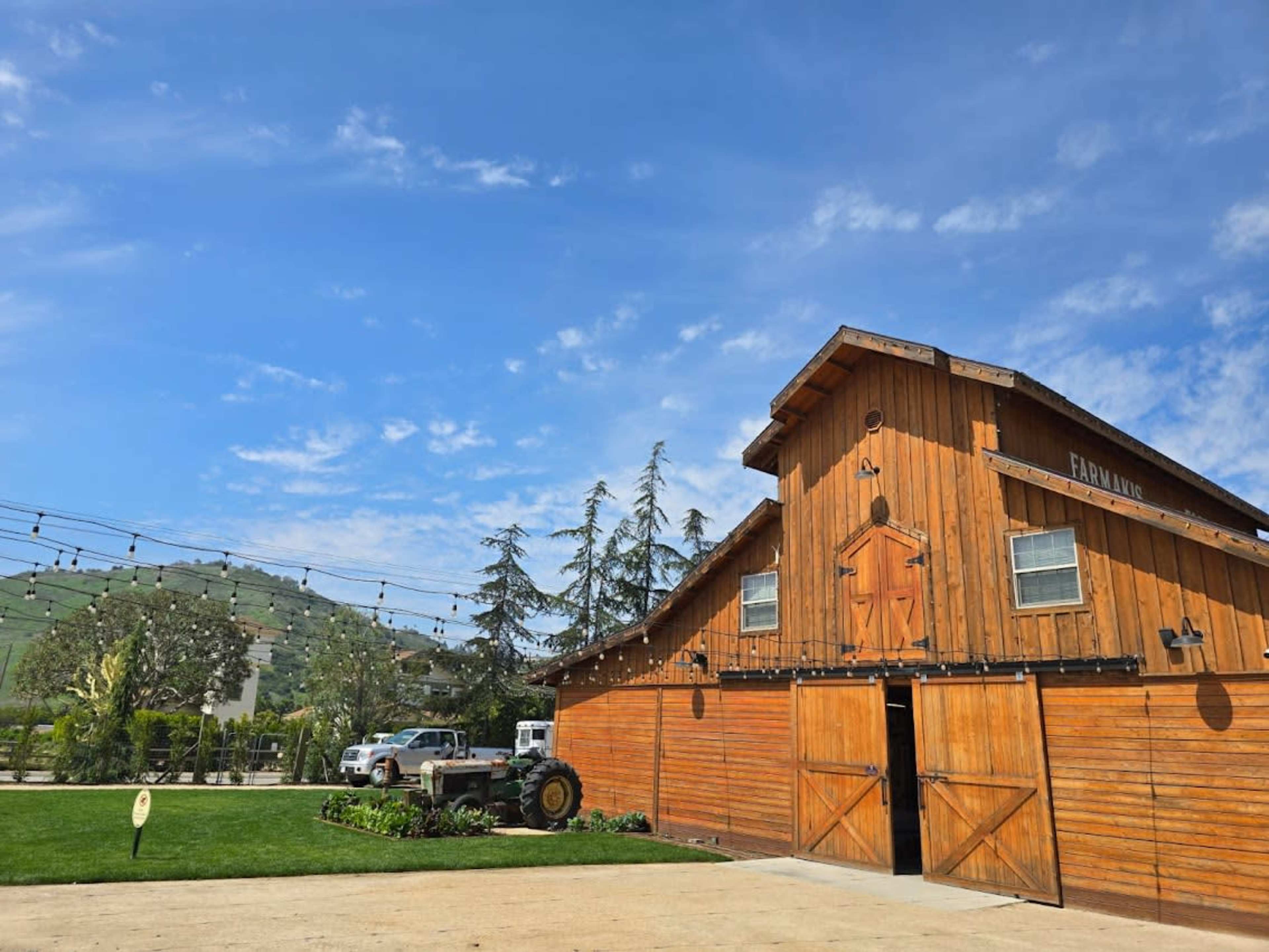 A wooden barn with open doors stands next to a tractor and a white truck, under a clear blue sky with scattered clouds.