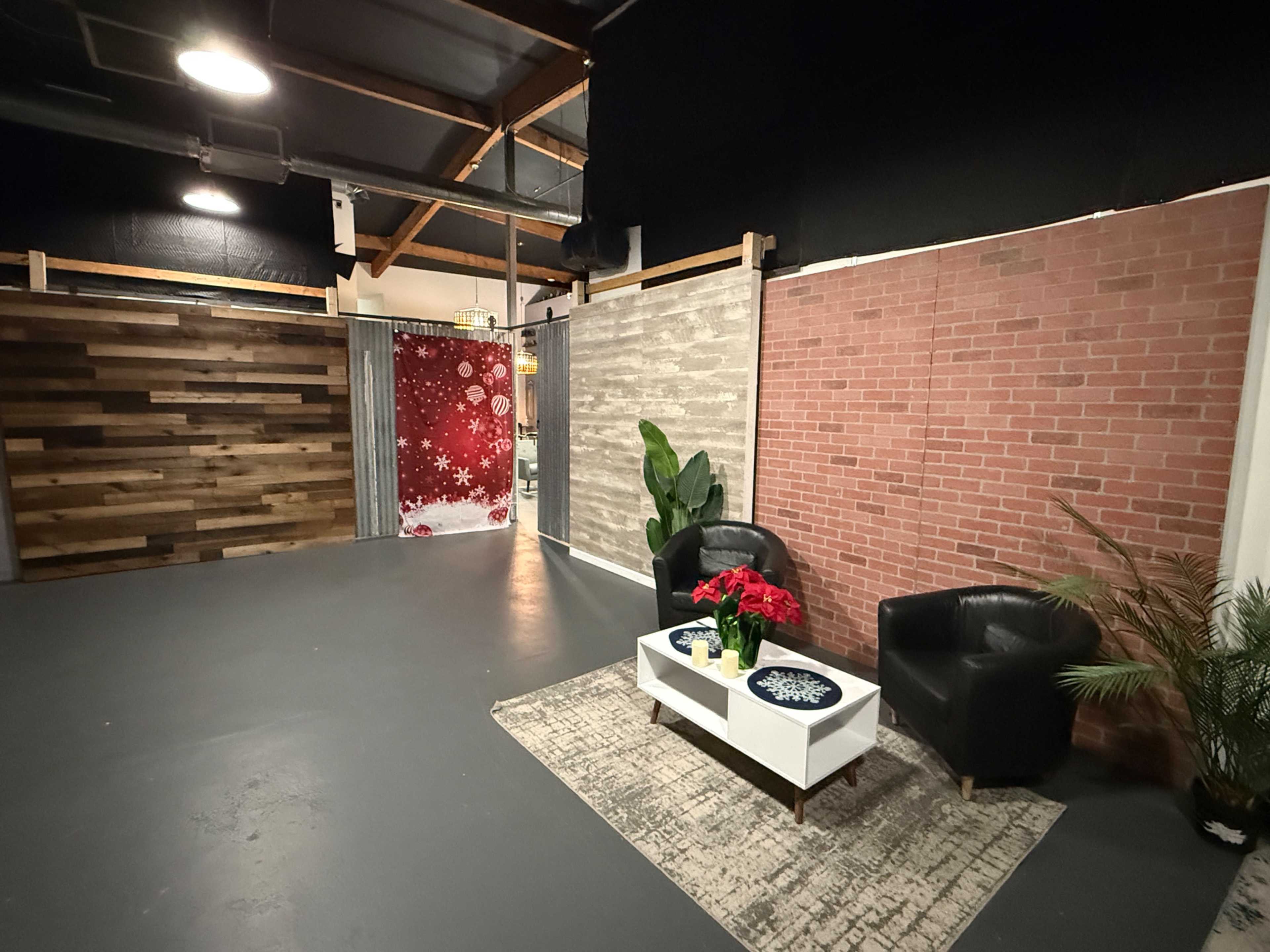The image shows a spacious interior with wooden and brick wall sections, two black armchairs, a white coffee table with a floral arrangement, and an area rug on a gray floor.