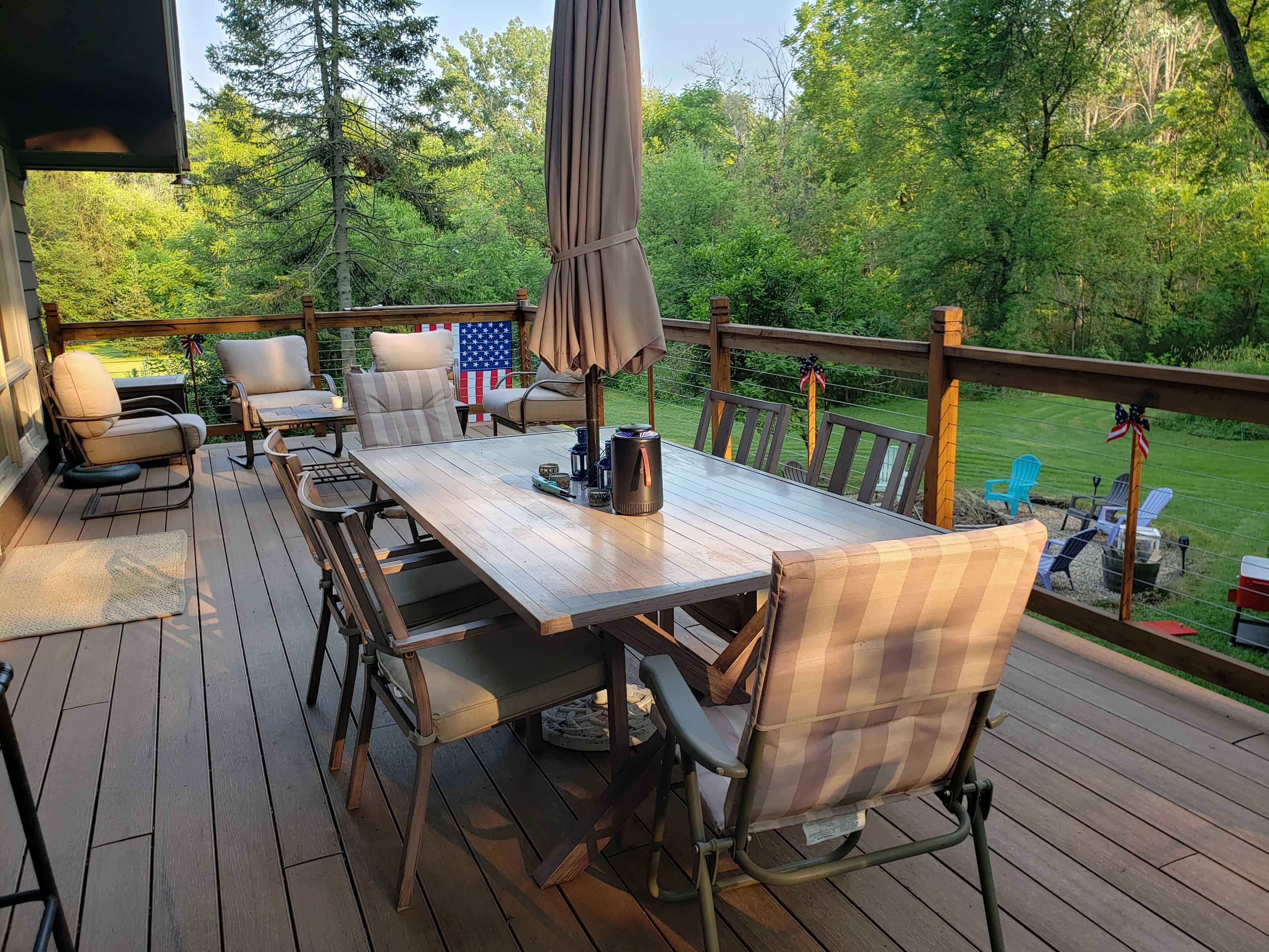 A wooden deck features a large table surrounded by multiple chairs, with an umbrella and decorative elements such as flags and outdoor seating visible in the background.