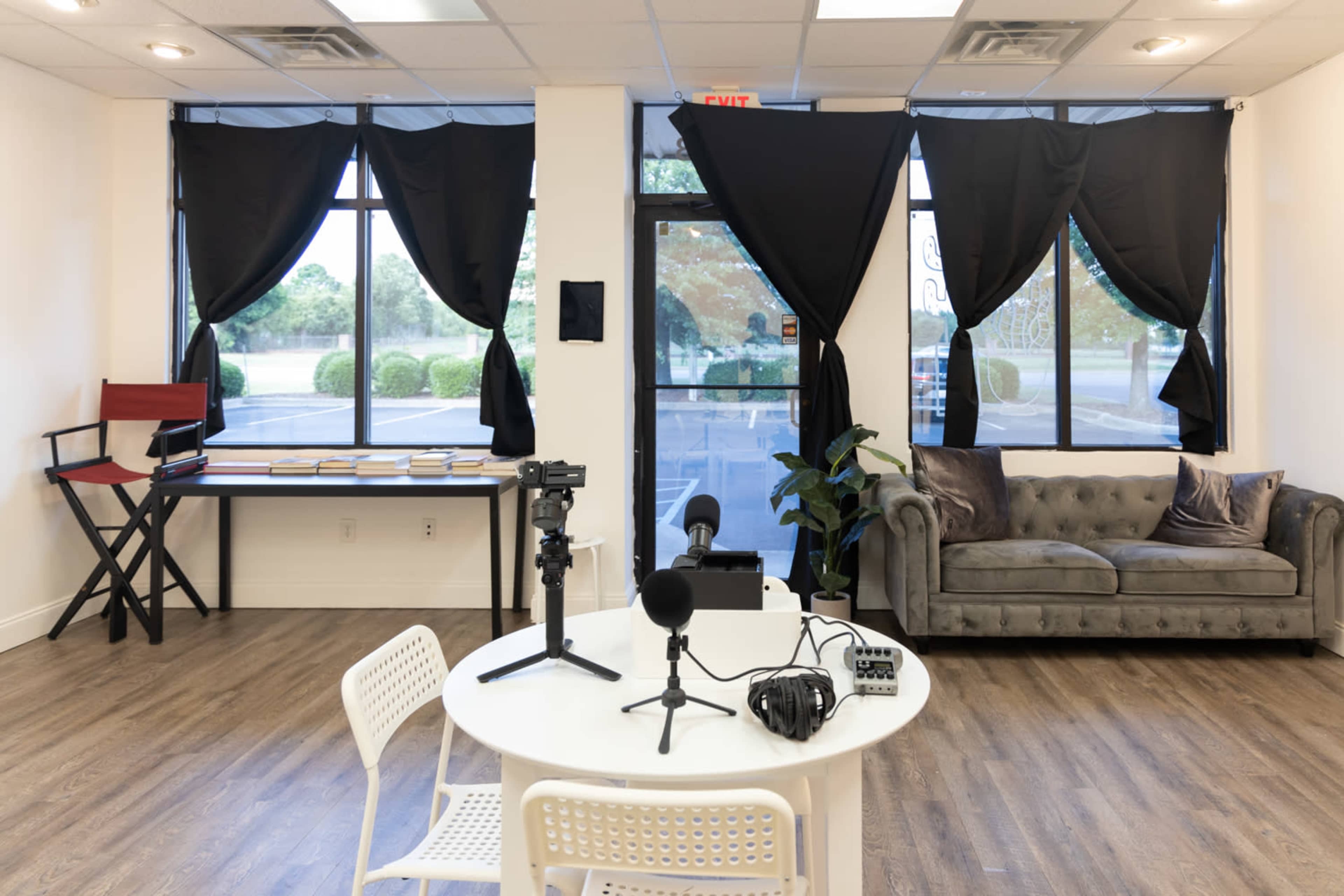 A studio space with a round table, microphones, a camera setup, and a gray couch near large windows draped with black curtains.