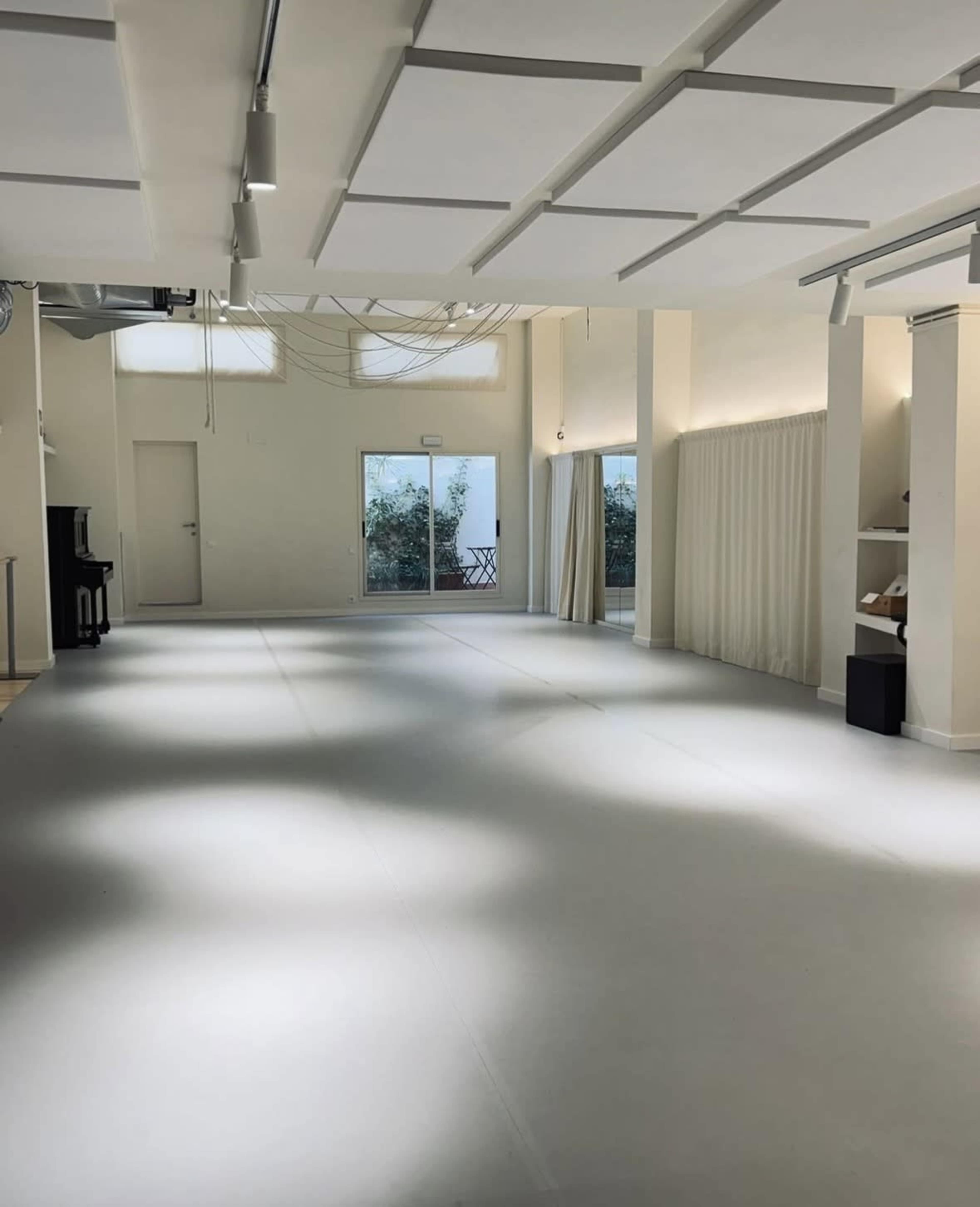 The image shows a spacious, well-lit studio with a large, empty floor, a piano in the corner, and soft natural light coming through windows.