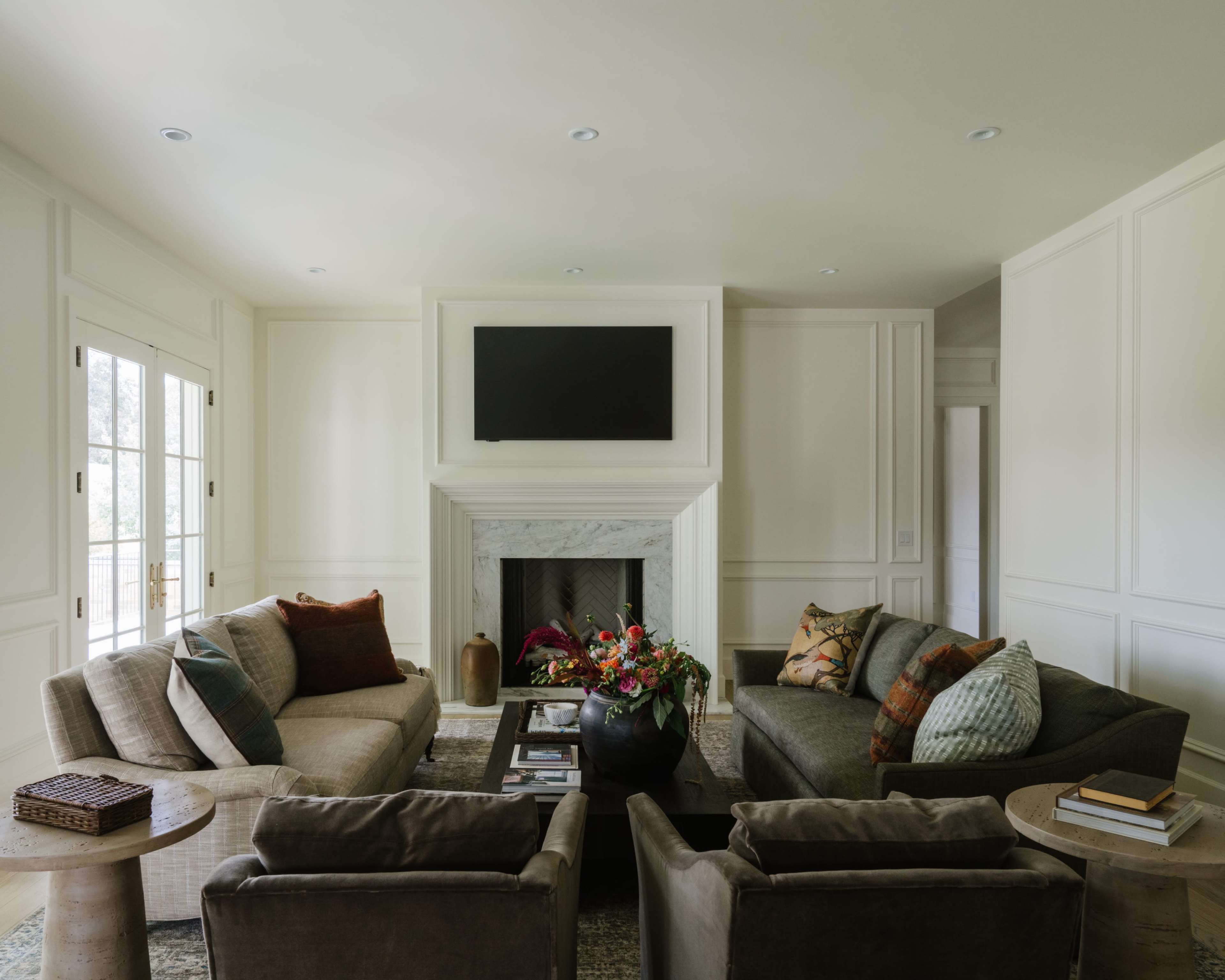 A modern living room features two sofas and two side tables arranged around a central coffee table, with a marble fireplace and a mounted television on the wall.