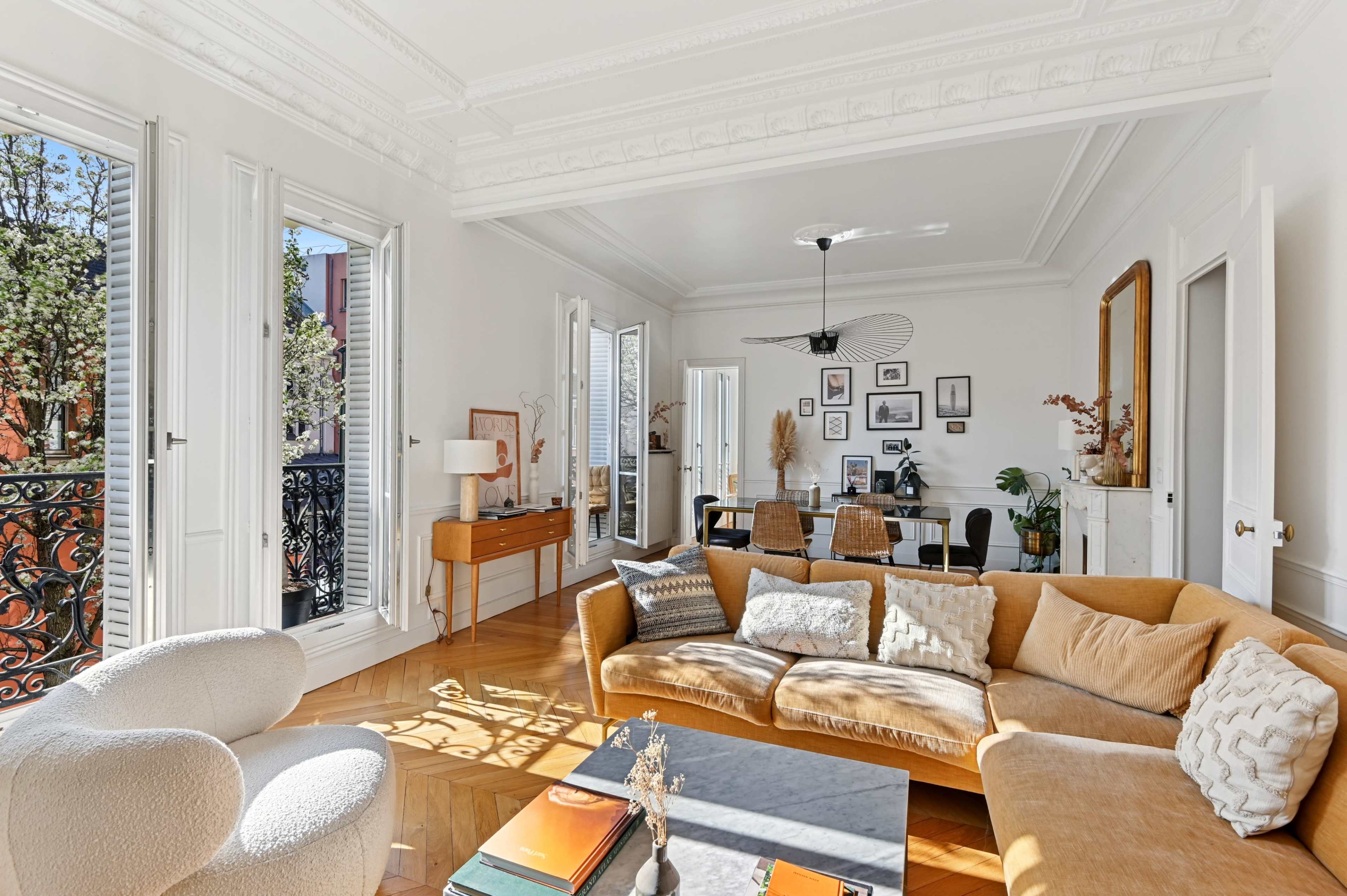 The image shows a bright, modern living room with a large sofa, a coffee table, and decorative furnishings, along with windows opening to a balcony.