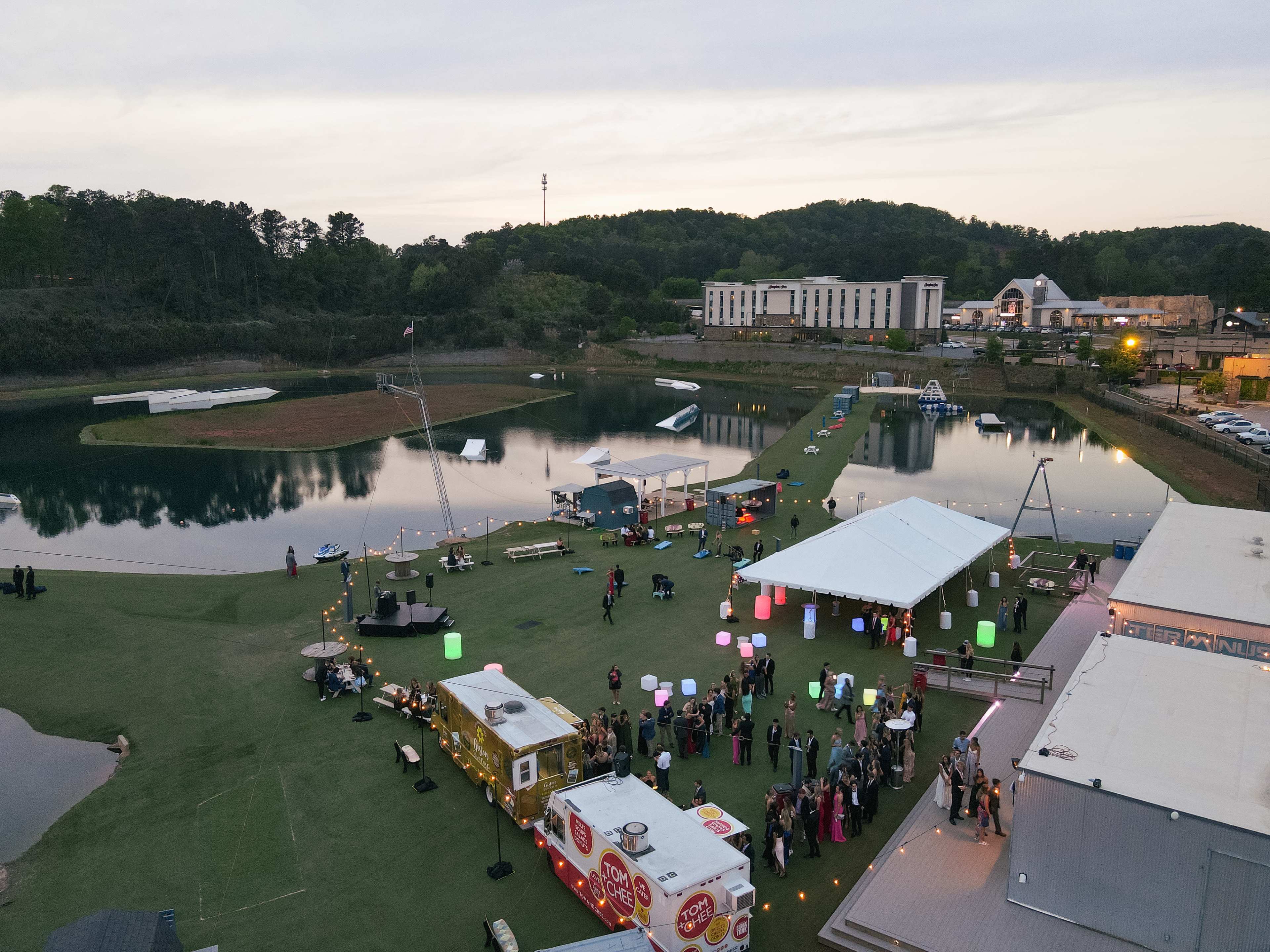 An aerial view of a lakeside event space featuring food trucks, colorful lanterns, and a gathering of people on a grassy area.