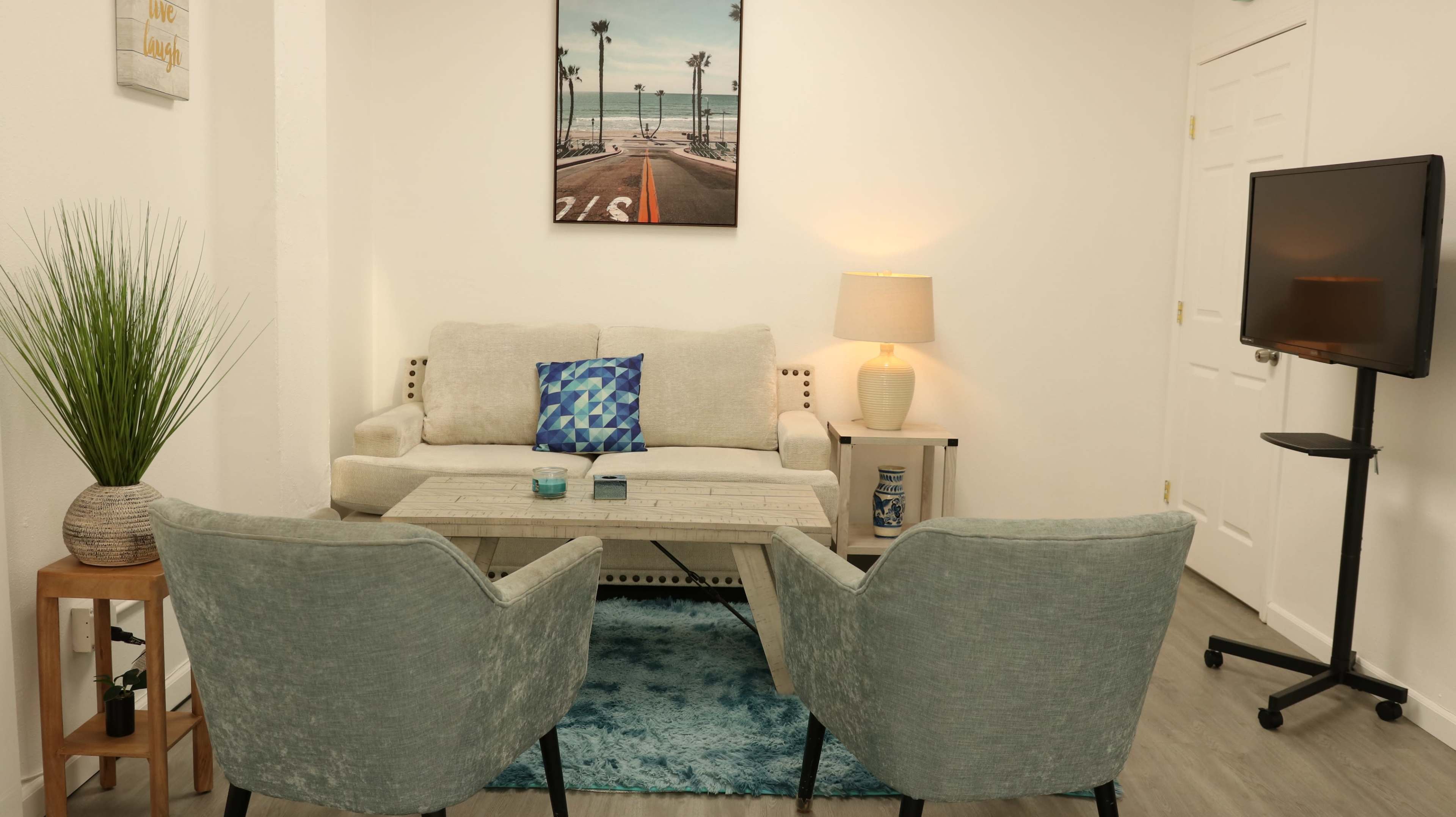 The image shows a well-decorated living area with a light-colored couch, two gray armchairs, a coffee table, and a wall-mounted TV, complemented by a palm tree-themed artwork and a plant in the corner.