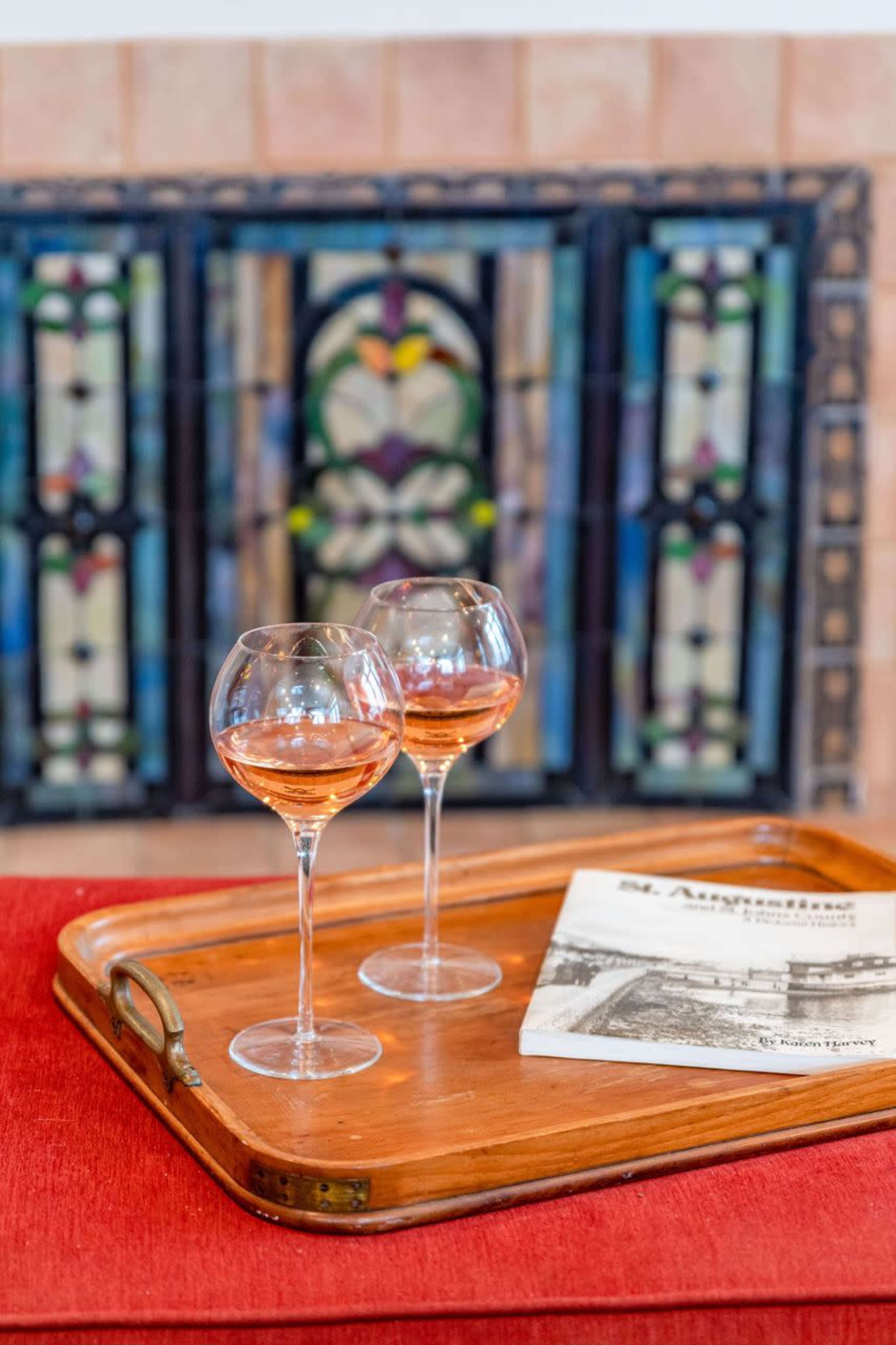 Two glasses of rosé wine are placed on a wooden tray next to a book, with a stained glass window in the blurred background.