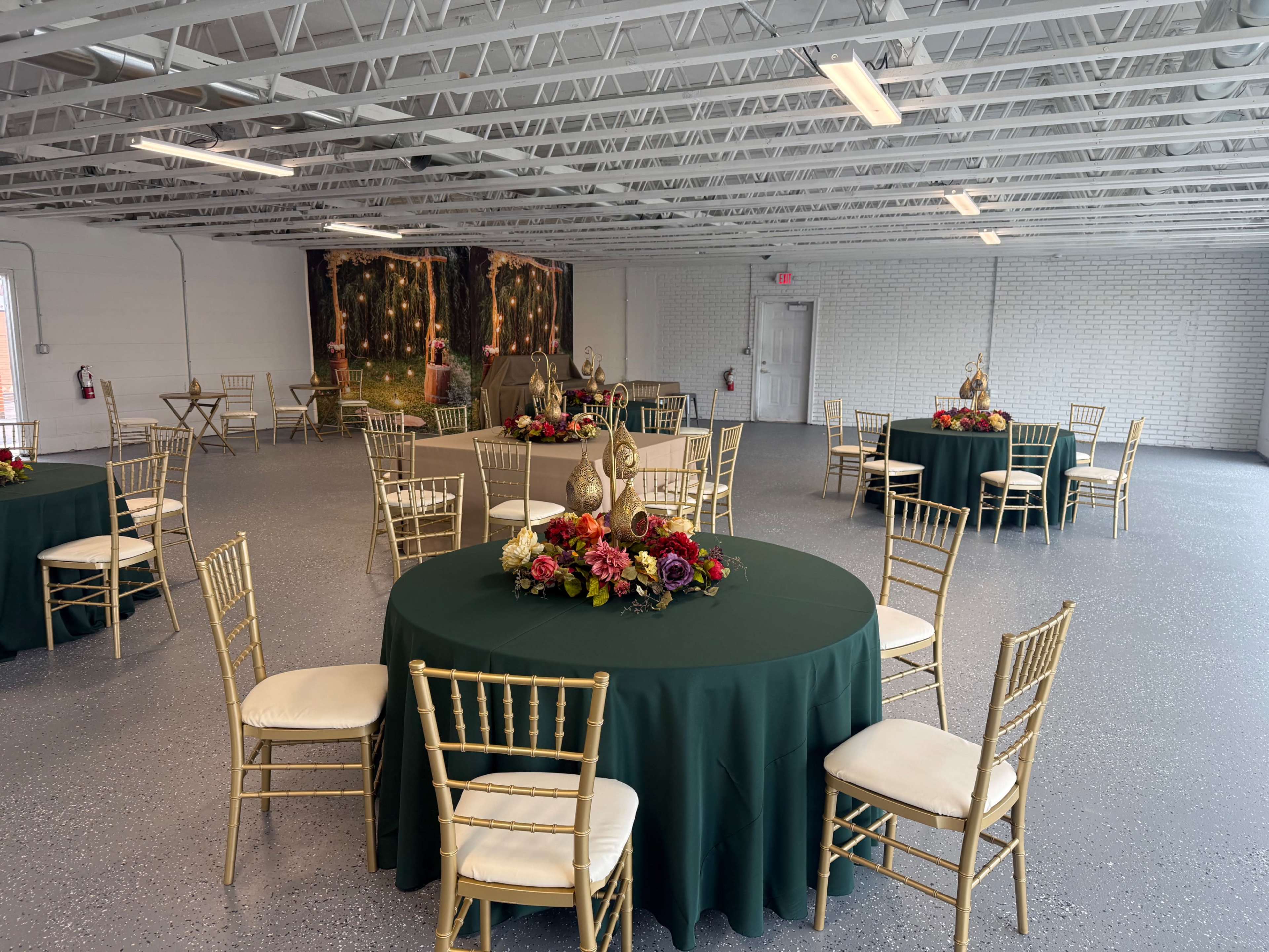 The image shows a spacious indoor event venue with several round tables covered in dark green tablecloths, arranged with floral centerpieces, and surrounded by gold Chiavari chairs.