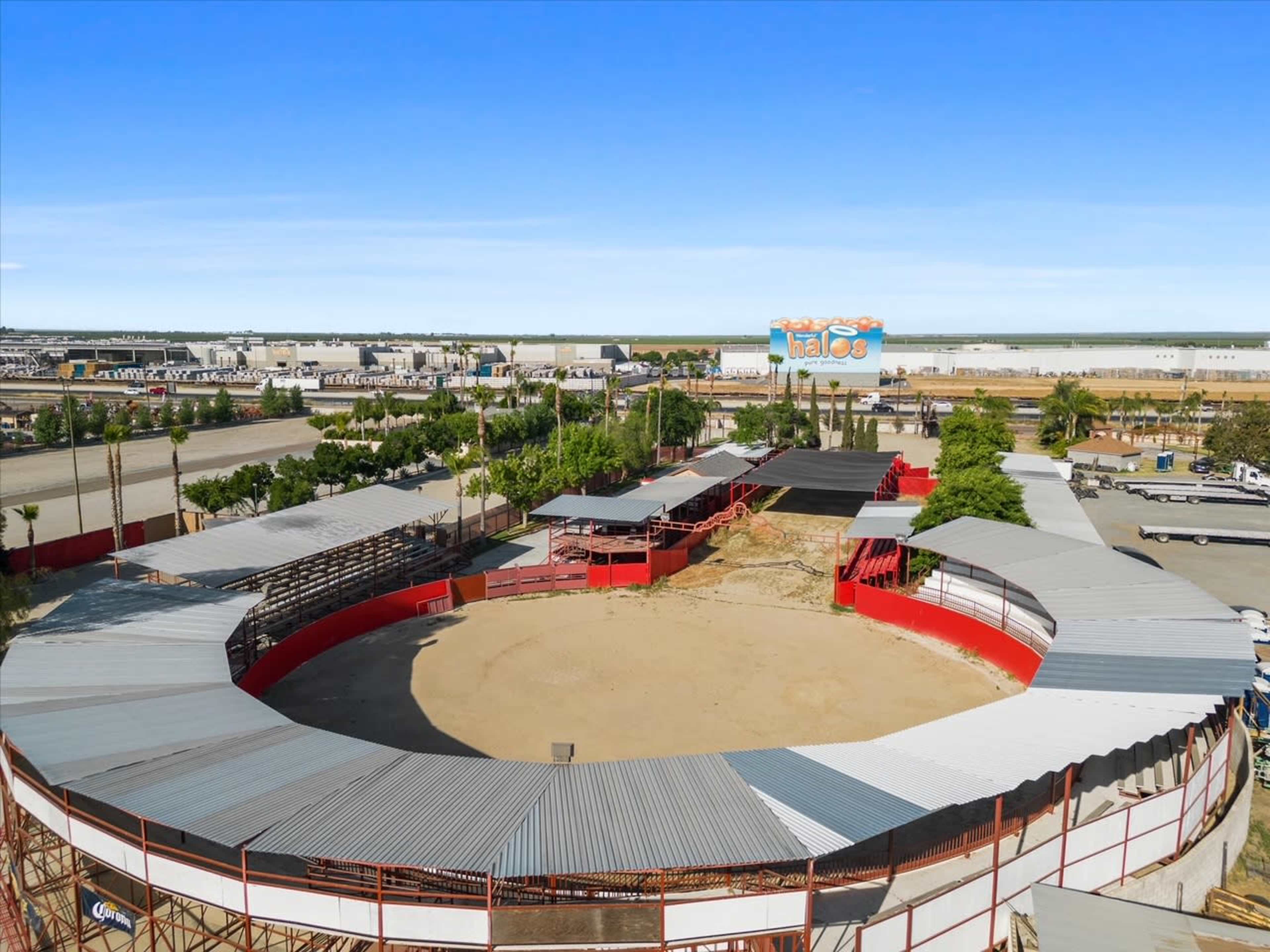 An aerial view of a circular arena surrounded by a red fence, situated in an industrial area with buildings and vehicles in the background.