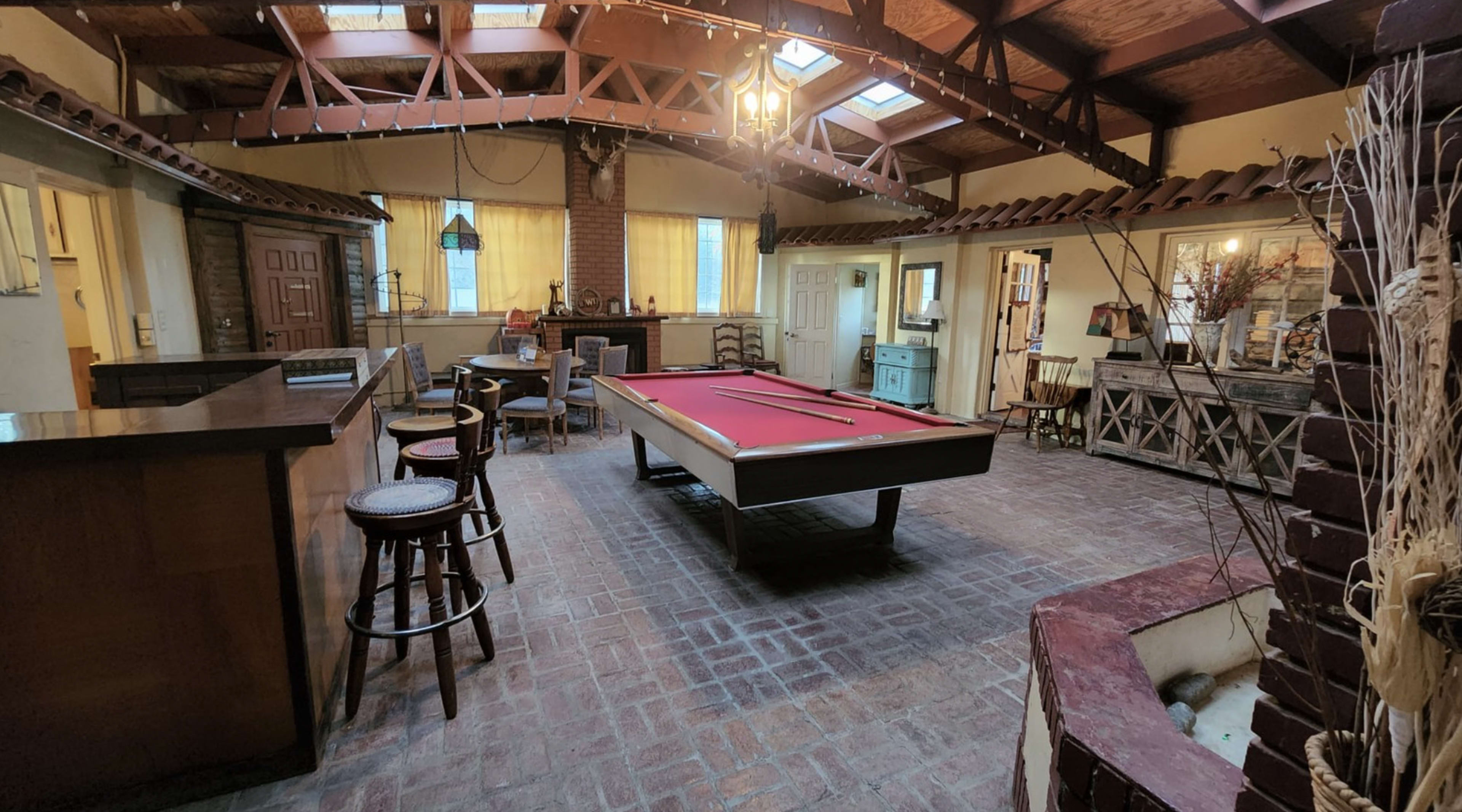 A spacious indoor room featuring a red billiard table centrally positioned, surrounded by seating areas and a rustic bar.