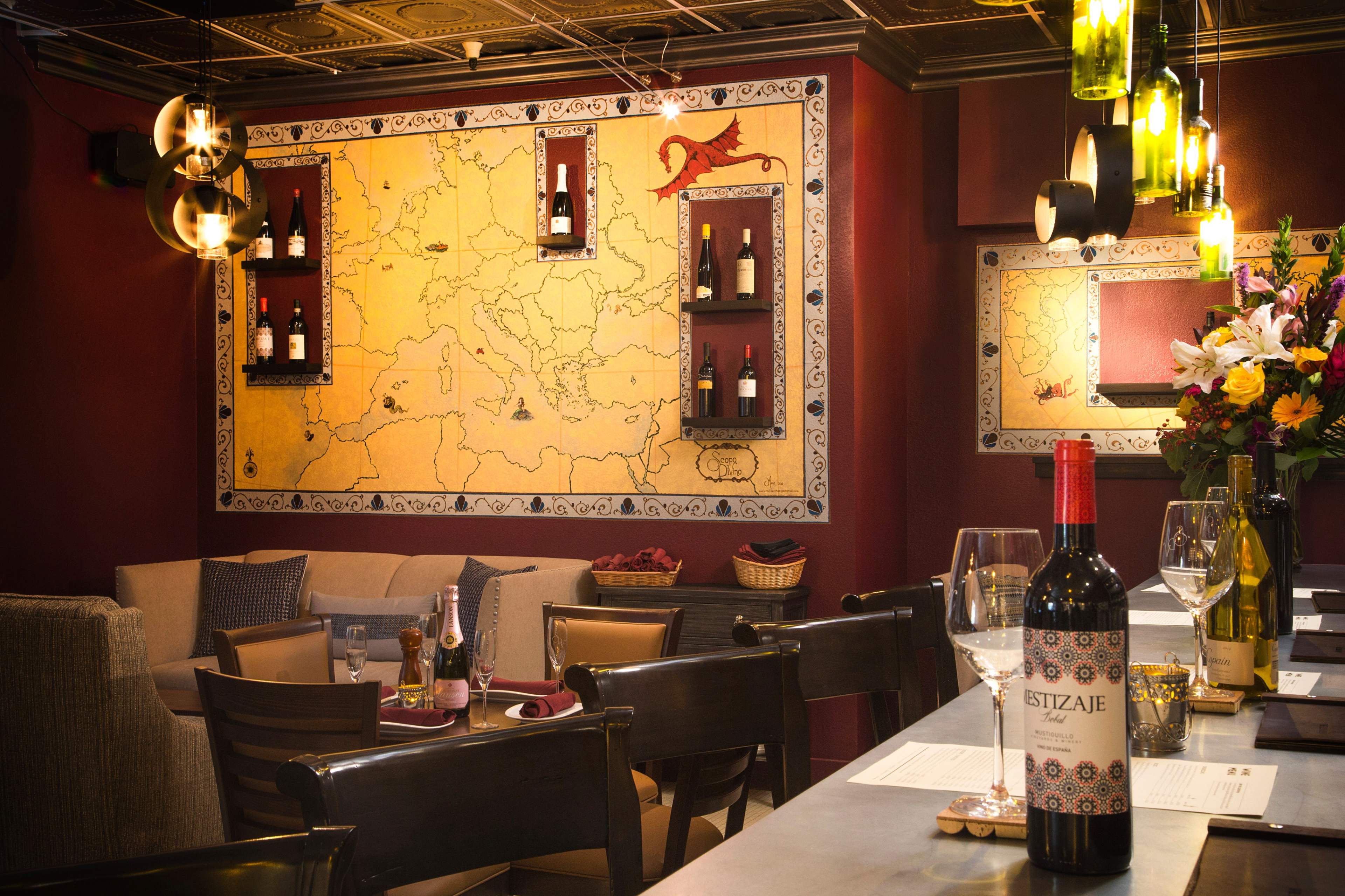 The interior of a restaurant features a decorative wall with a large map design and shelves displaying wine bottles, complemented by elegant table settings.