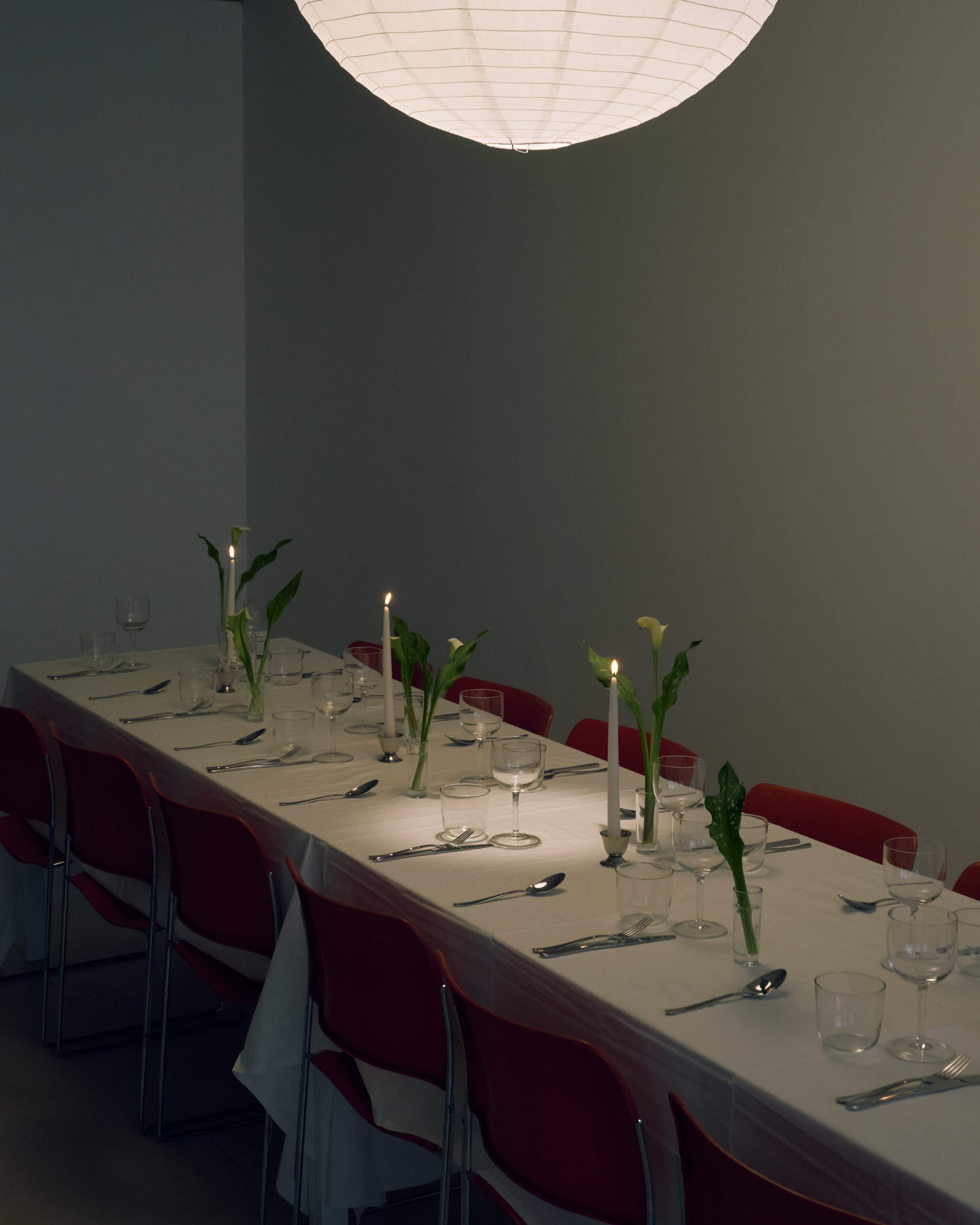 A long dining table is set with an ornate white tablecloth, glassware, and candles, with red chairs positioned around it and a paper lantern hanging above.