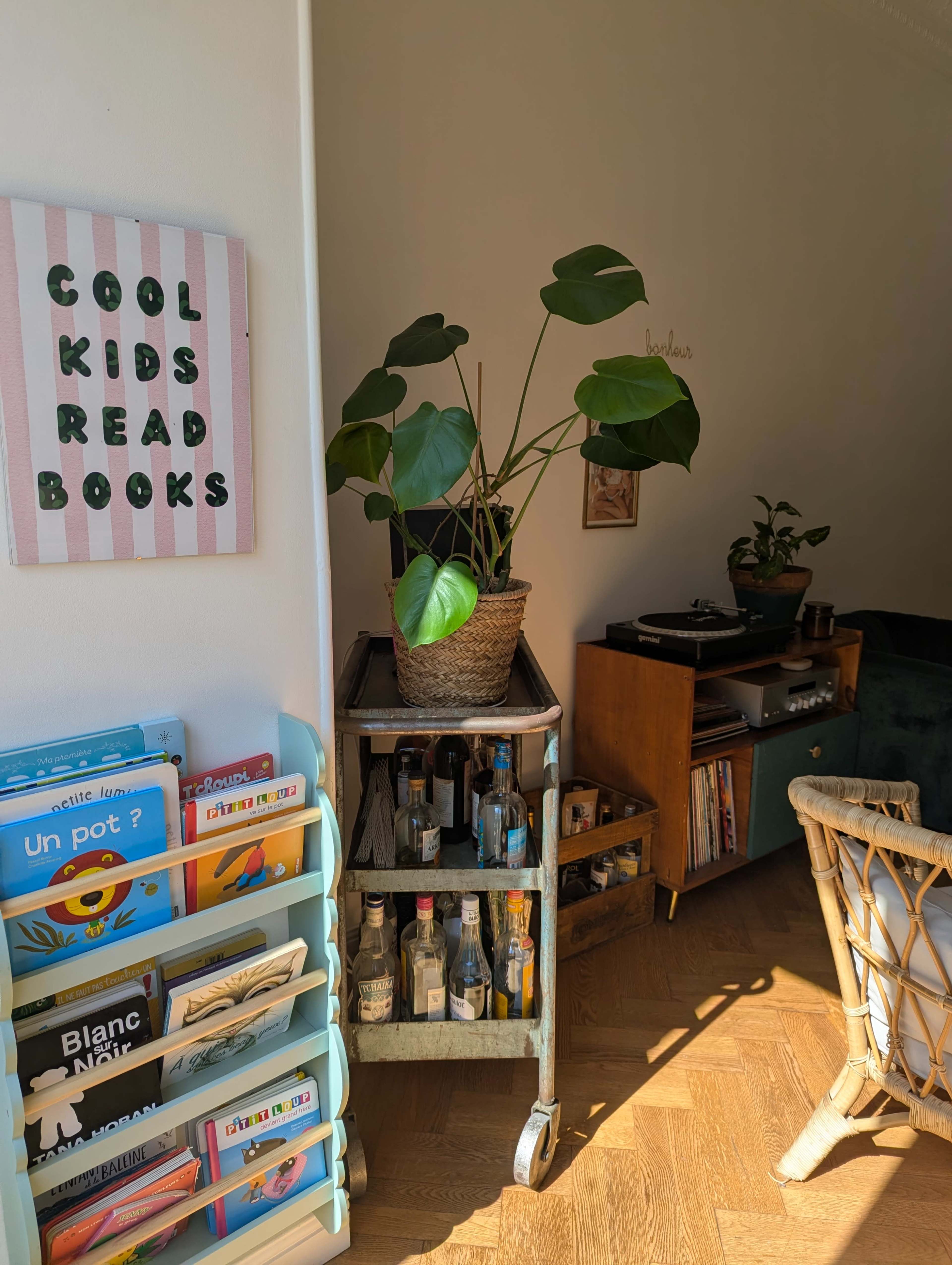 The image shows a cozy corner of a room featuring a plant on a wheeled cart stocked with bottles, a bookshelf filled with children's books, and a vintage record player with a collection of vinyl records.