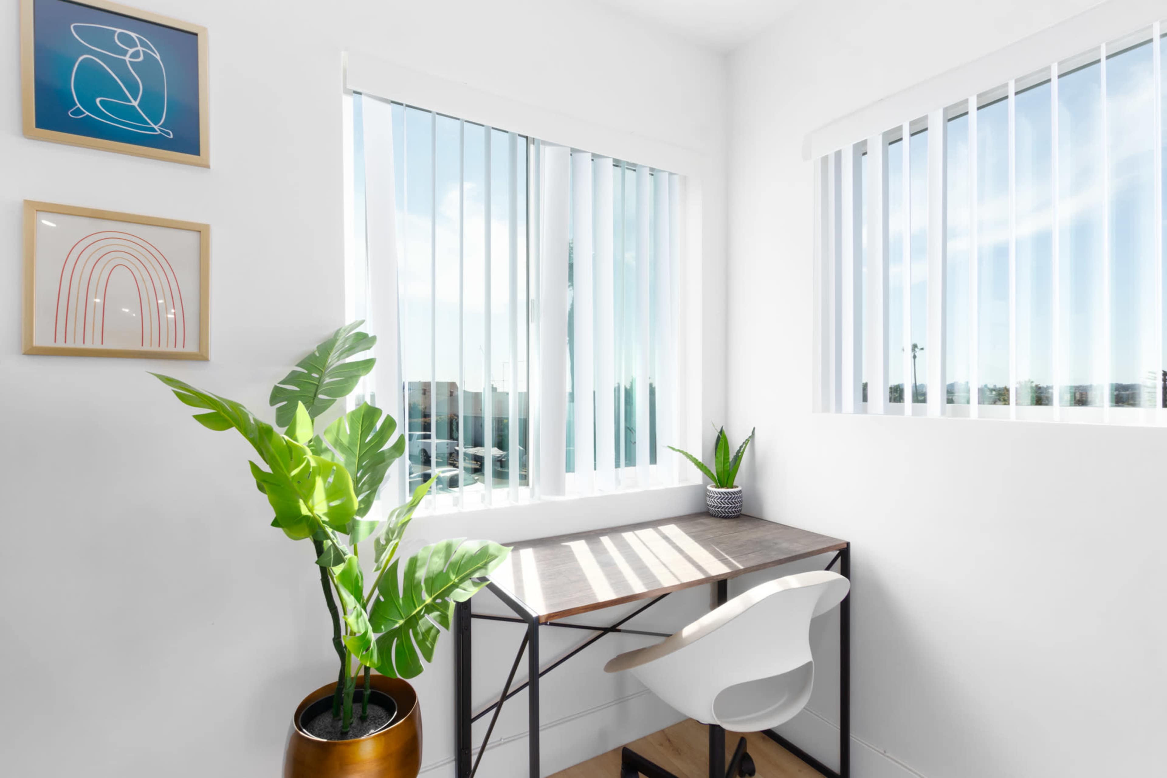 A modern workspace features a desk and chair by a window, with a potted plant and decorative wall art in a bright room.