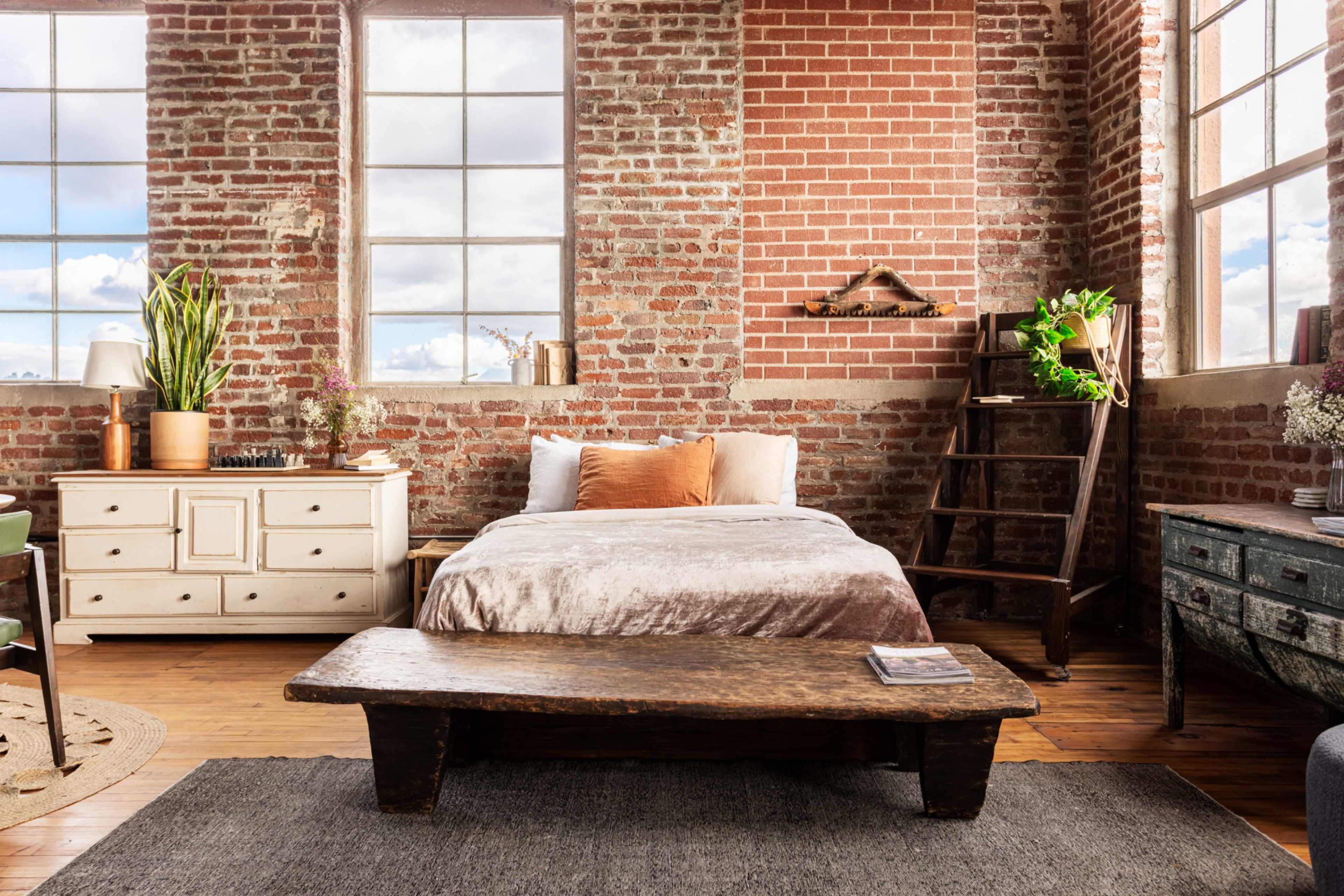 A cozy bedroom featuring a bed against a brick wall, a wooden coffee table, a dresser, and large windows that allow natural light to illuminate the space.
