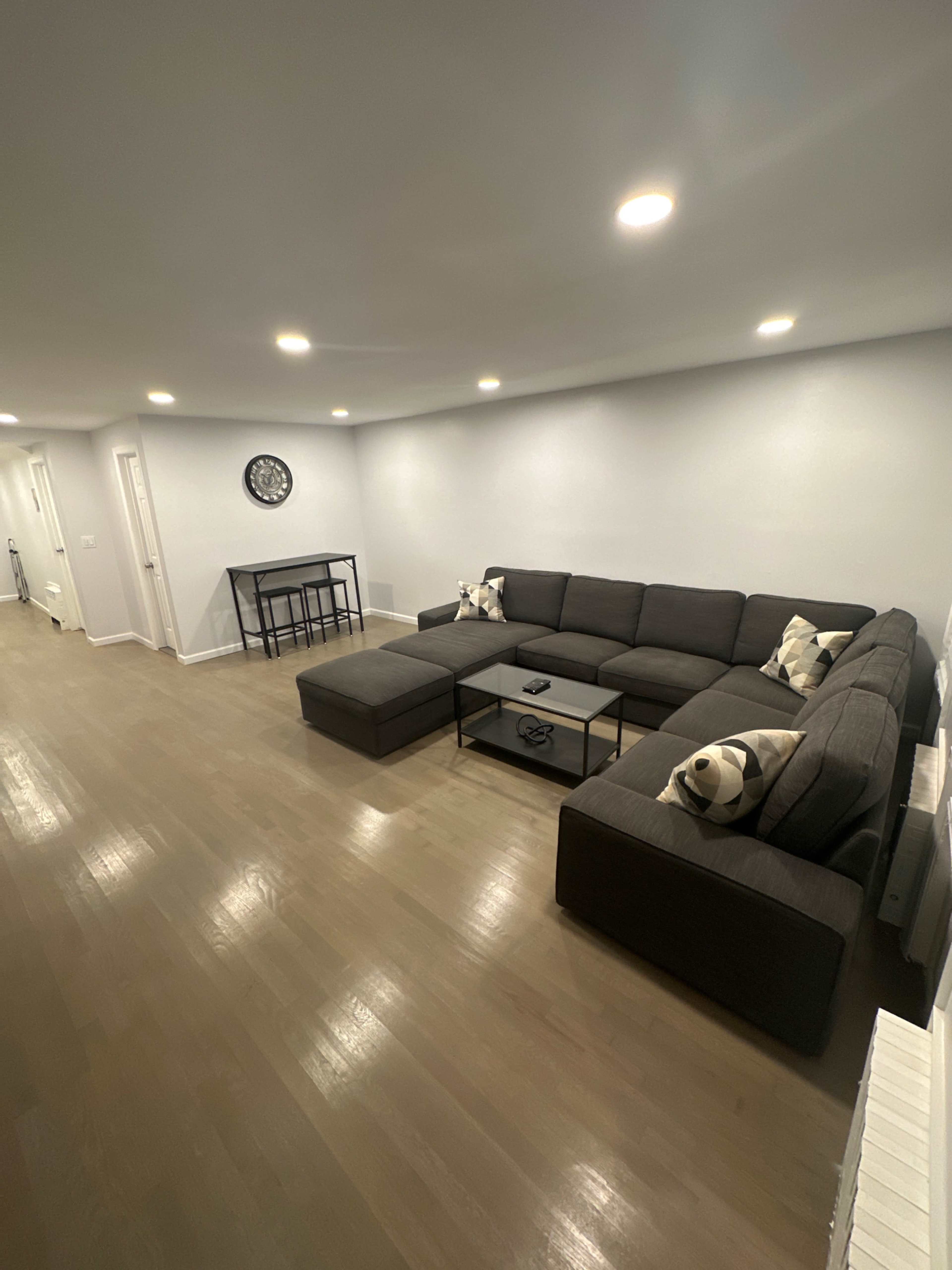 The image shows a spacious living room featuring a large gray sectional sofa with decorative pillows and a coffee table, along with a small dining table in the background.