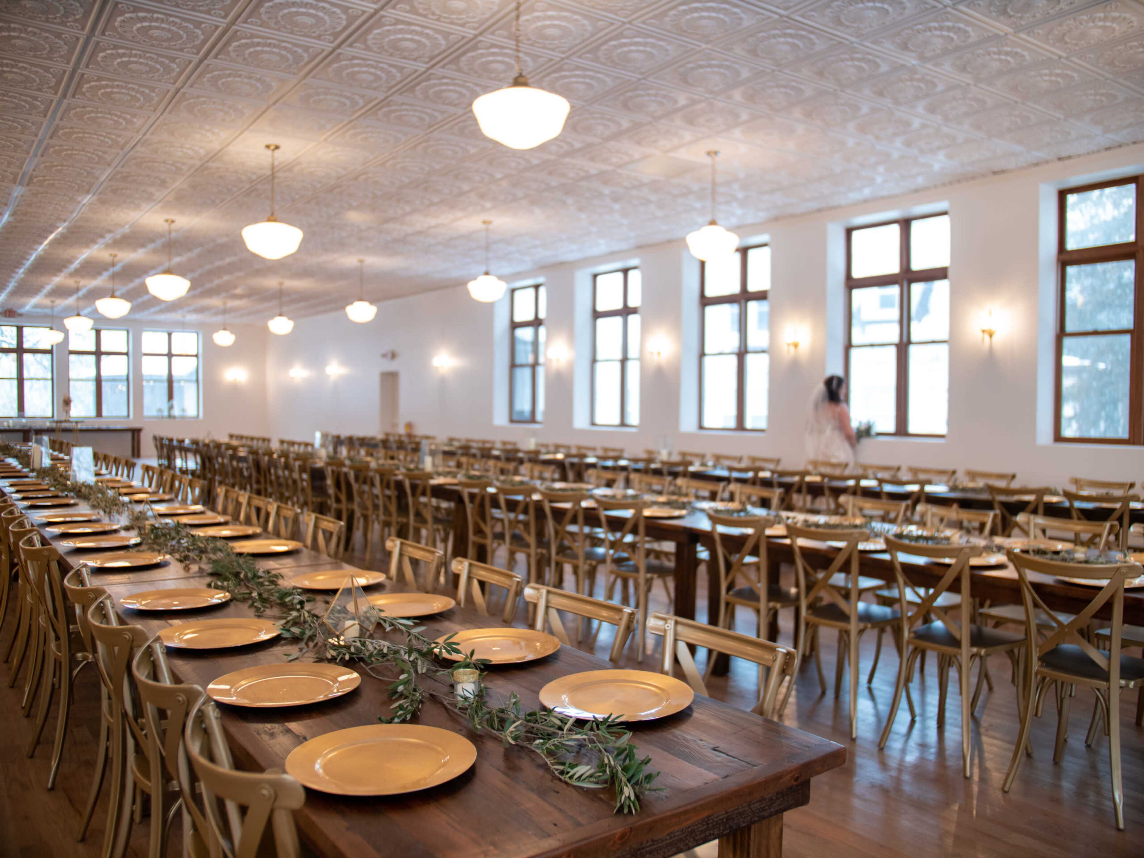 A spacious banquet hall is set with long tables adorned with gold plates and greenery, illuminated by overhead lights and large windows.