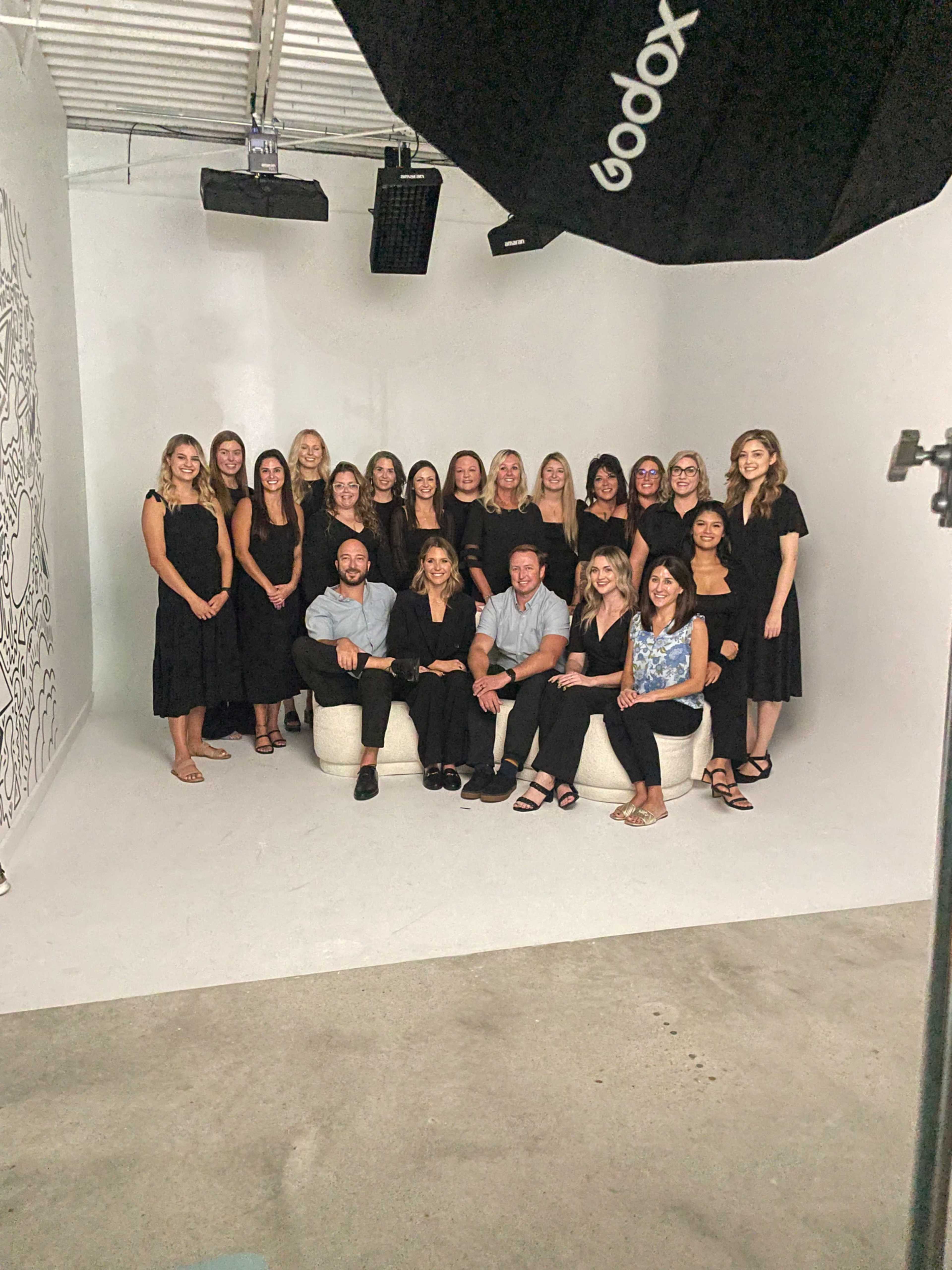 A group of 20 individuals, dressed in black and seated on light-colored furniture, poses for a photo in a well-lit studio setting.