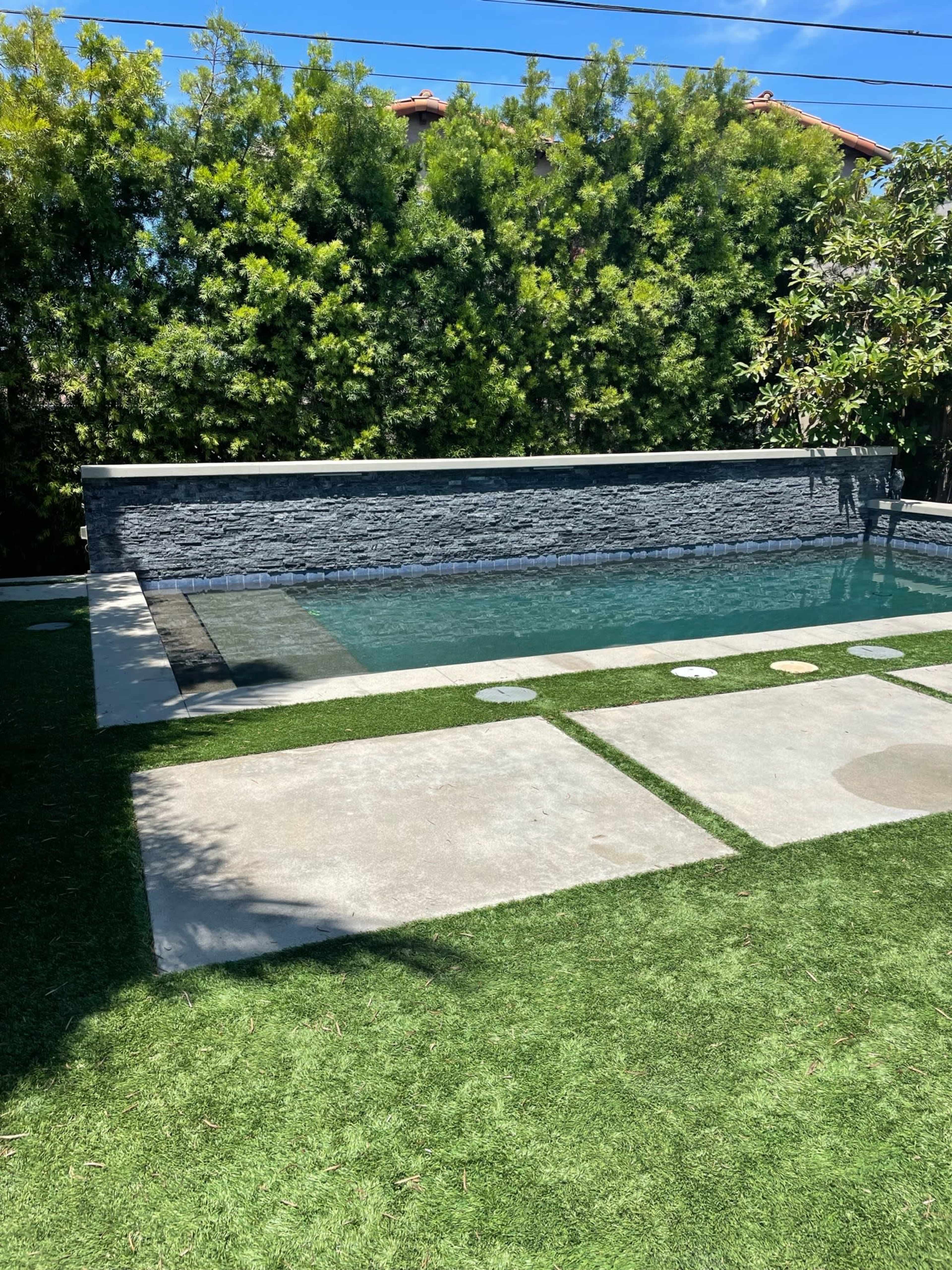 A modern backyard featuring a rectangular pool with a stone wall backdrop and a grassy area bordered by concrete pathways.
