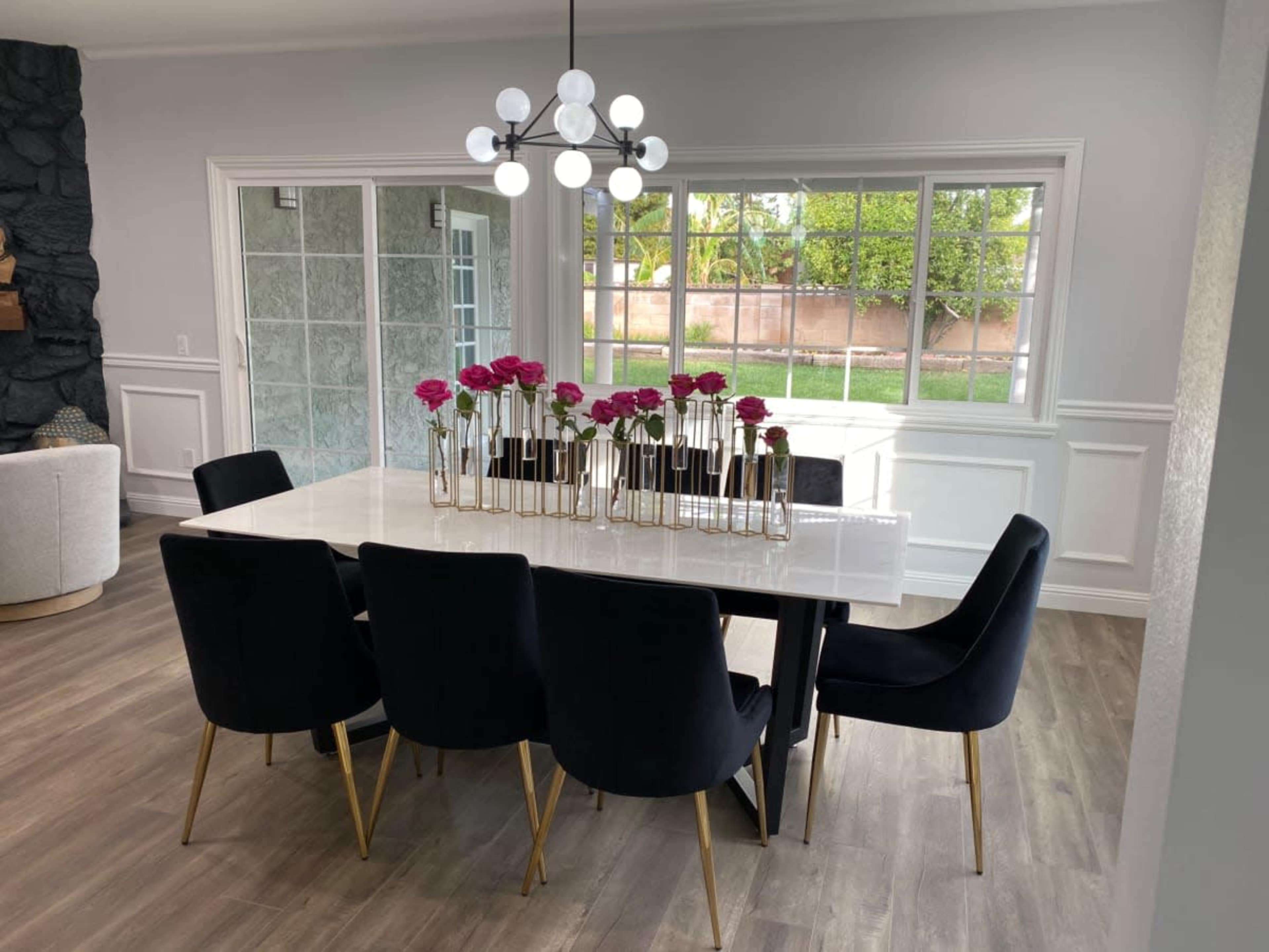 A modern dining room features a large white table surrounded by black chairs, with a decorative centerpiece of pink flowers and a contemporary light fixture overhead.