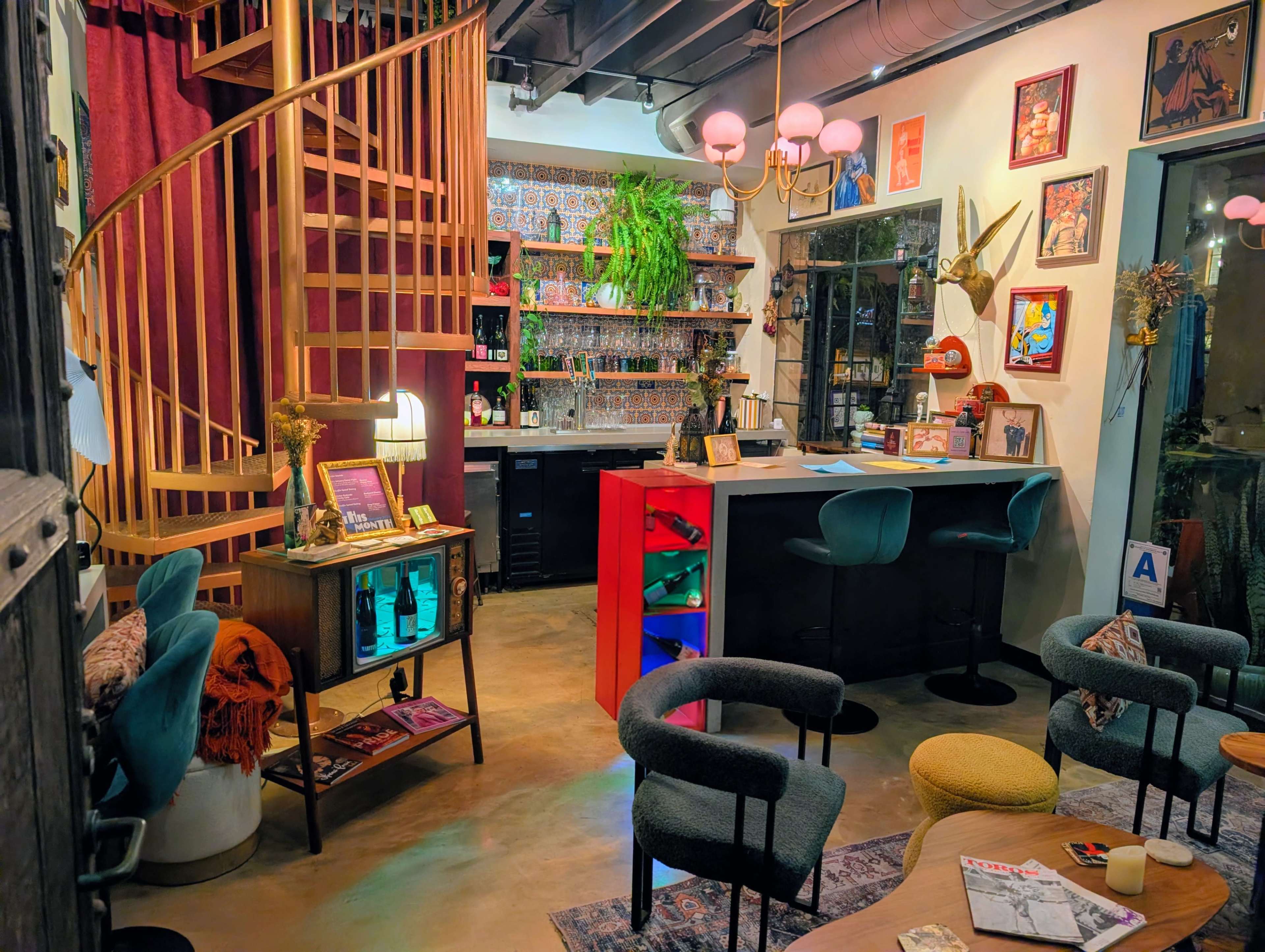 The image shows a cozy, eclectic café interior with a spiral staircase, a well-stocked bar, and various seating areas decorated with plants and artwork.
