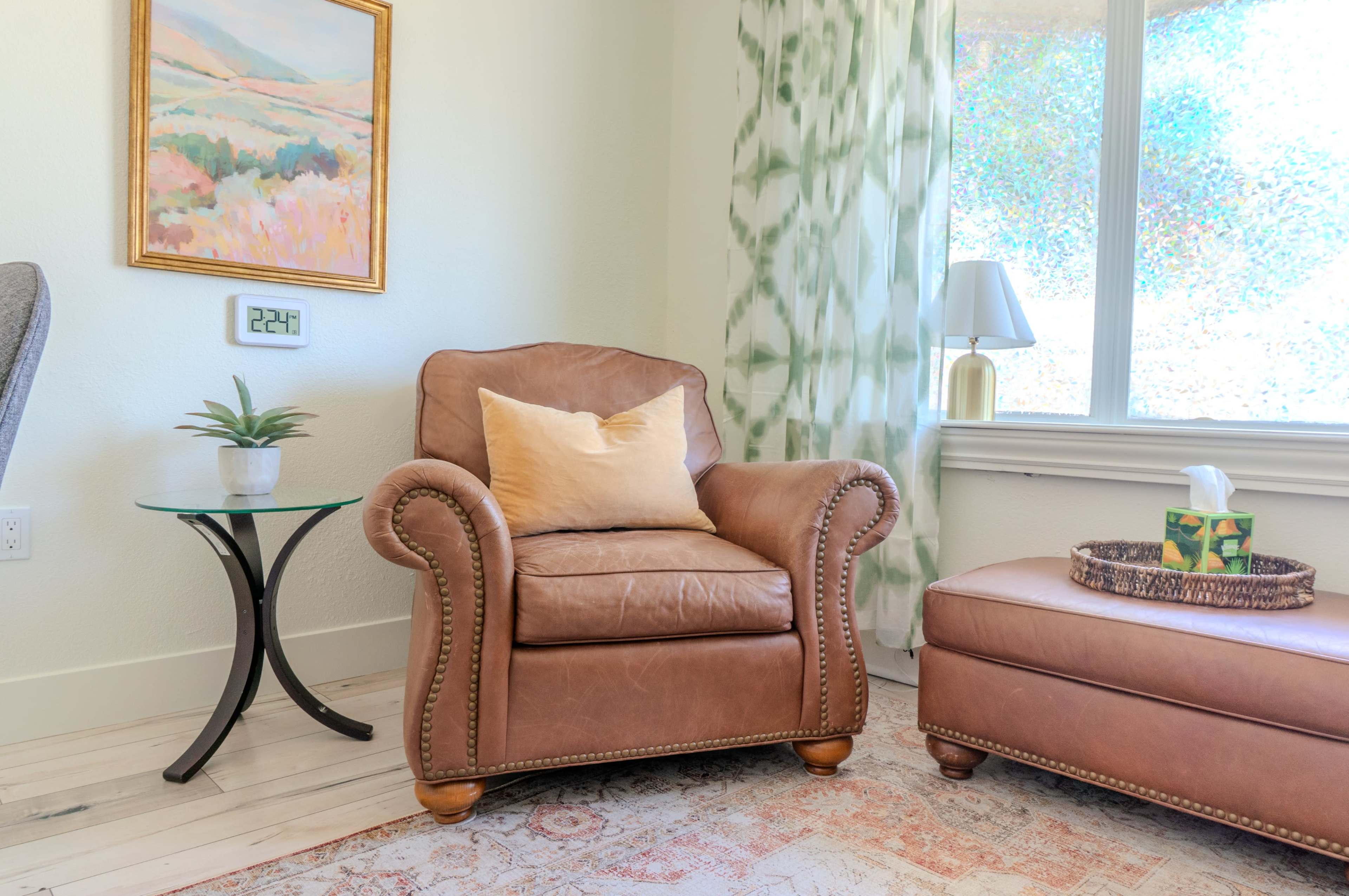 A leather armchair with a pillow sits next to a small glass side table holding a decorative lamp and a tissue box, in a well-lit room with a window and patterned curtains.
