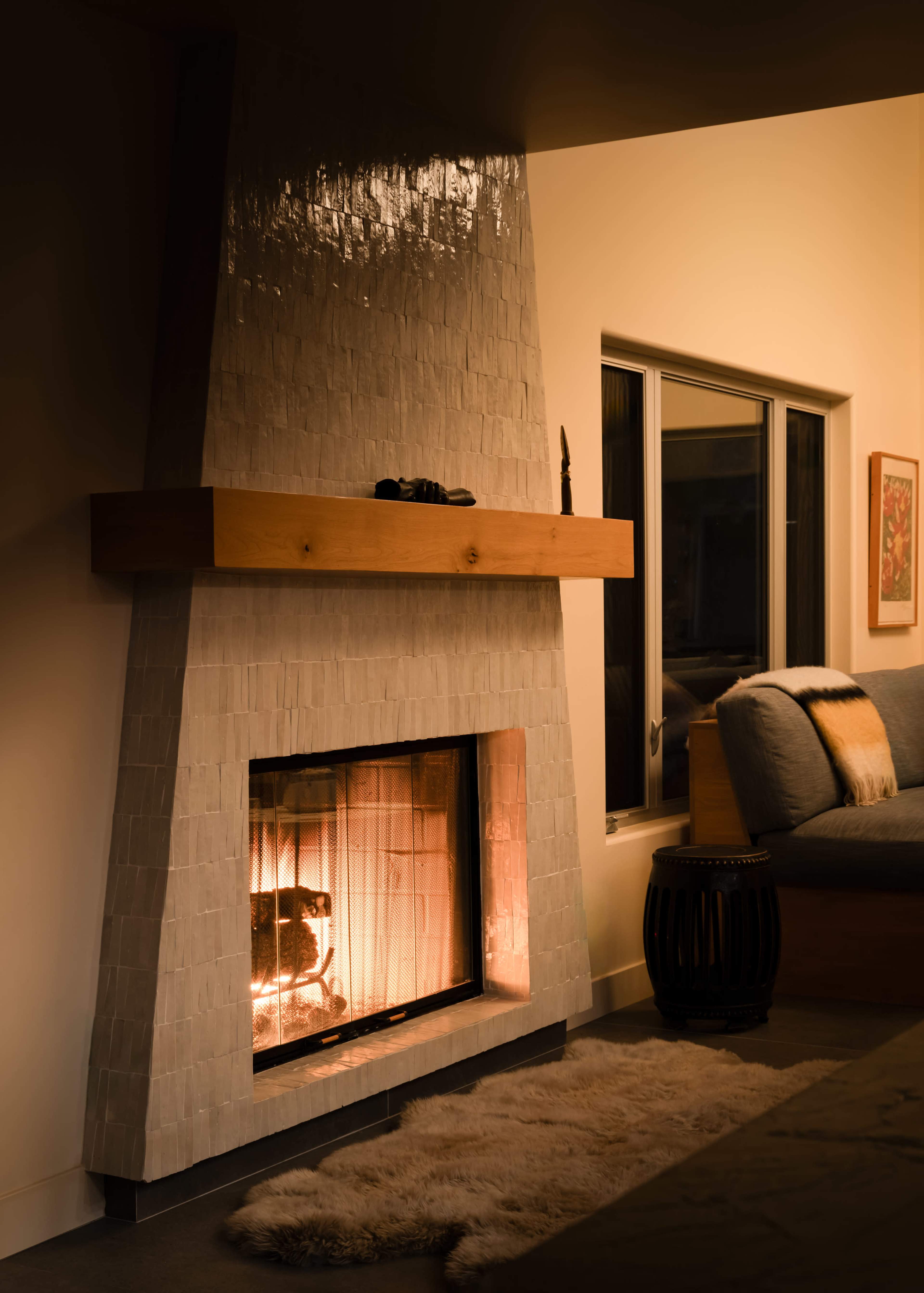 A modern fireplace with a tiled facade and wooden mantel is situated in a cozy living room, featuring a soft rug and a window beside it.