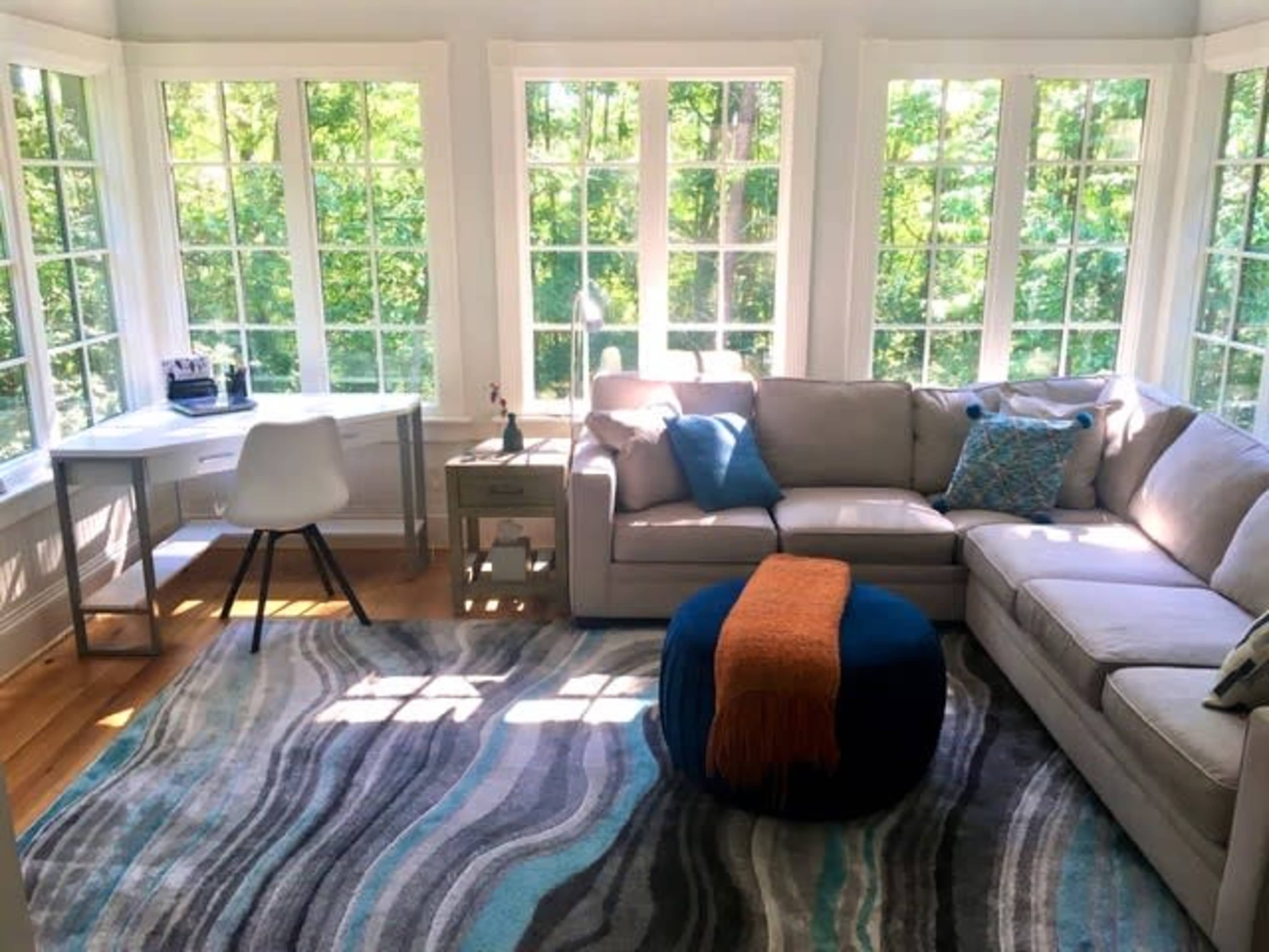The image shows a bright living room with large windows surrounded by greenery, featuring a sectional sofa, a round ottoman, and a desk set up against the wall.